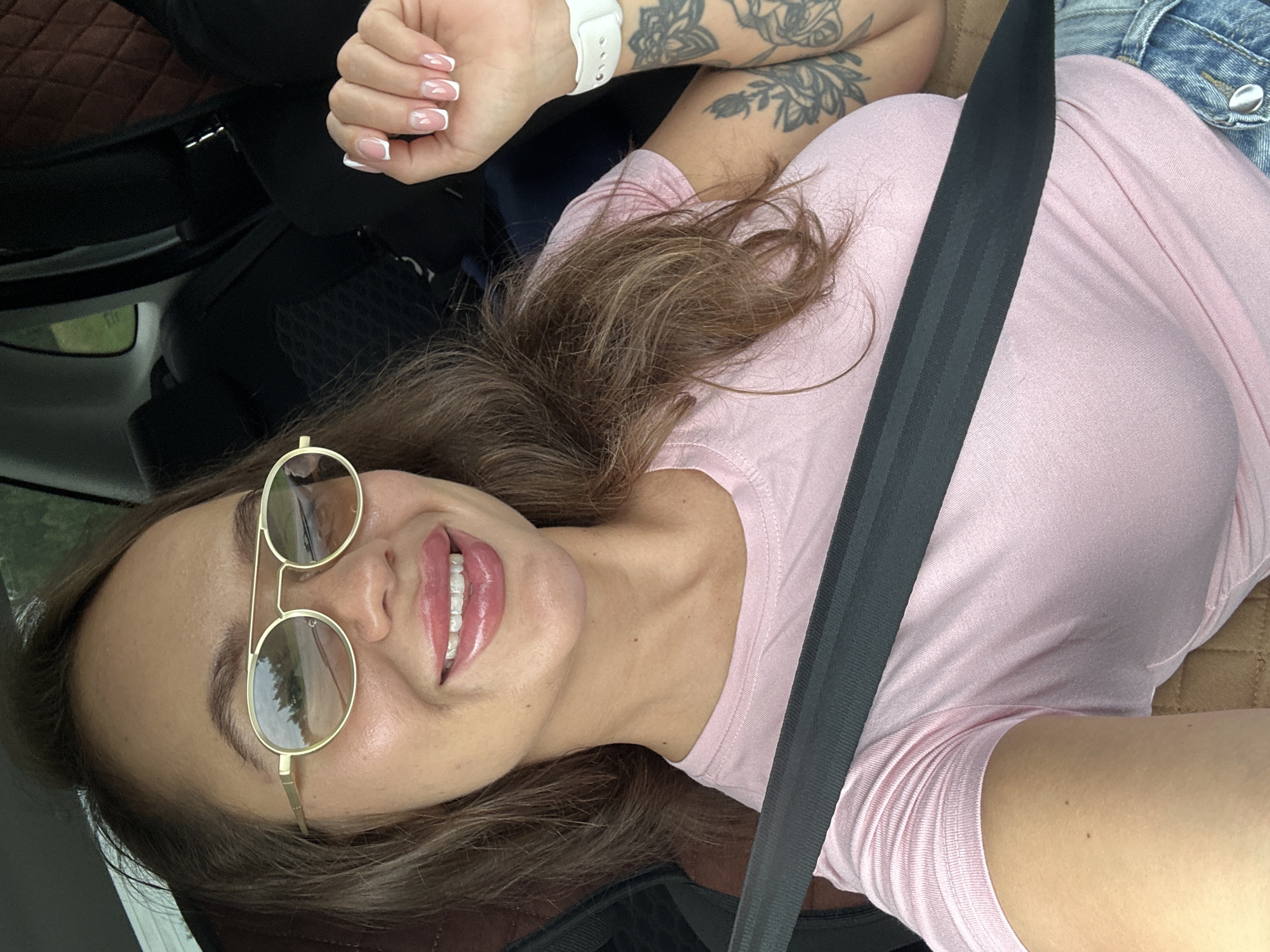 A woman with long brown hair, wearing sunglasses, a pink t-shirt, and blue jeans, is seen from the chest up, inside a car. She is wearing a seatbelt and has tattoos on her left arm. Her fingernails are painted with French tips. The background shows car seats and windows. The image appears to have been taken on a cloudy day. The location is specified as Uvel'skiy, Russia.