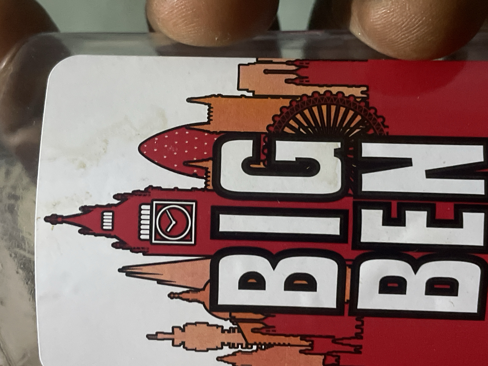 The image shows a close-up of a sticker on a jar. The sticker features a stylized illustration of the London skyline, including Big Ben and the London Eye. The words "BIG BEN" are printed in large white letters with black outlines across the lower portion of the sticker. A person's fingers are visible on the right side of the image, partially obscuring the jar. There are no discernible elements in the image that would indicate a specific location in Jos, Nigeria.