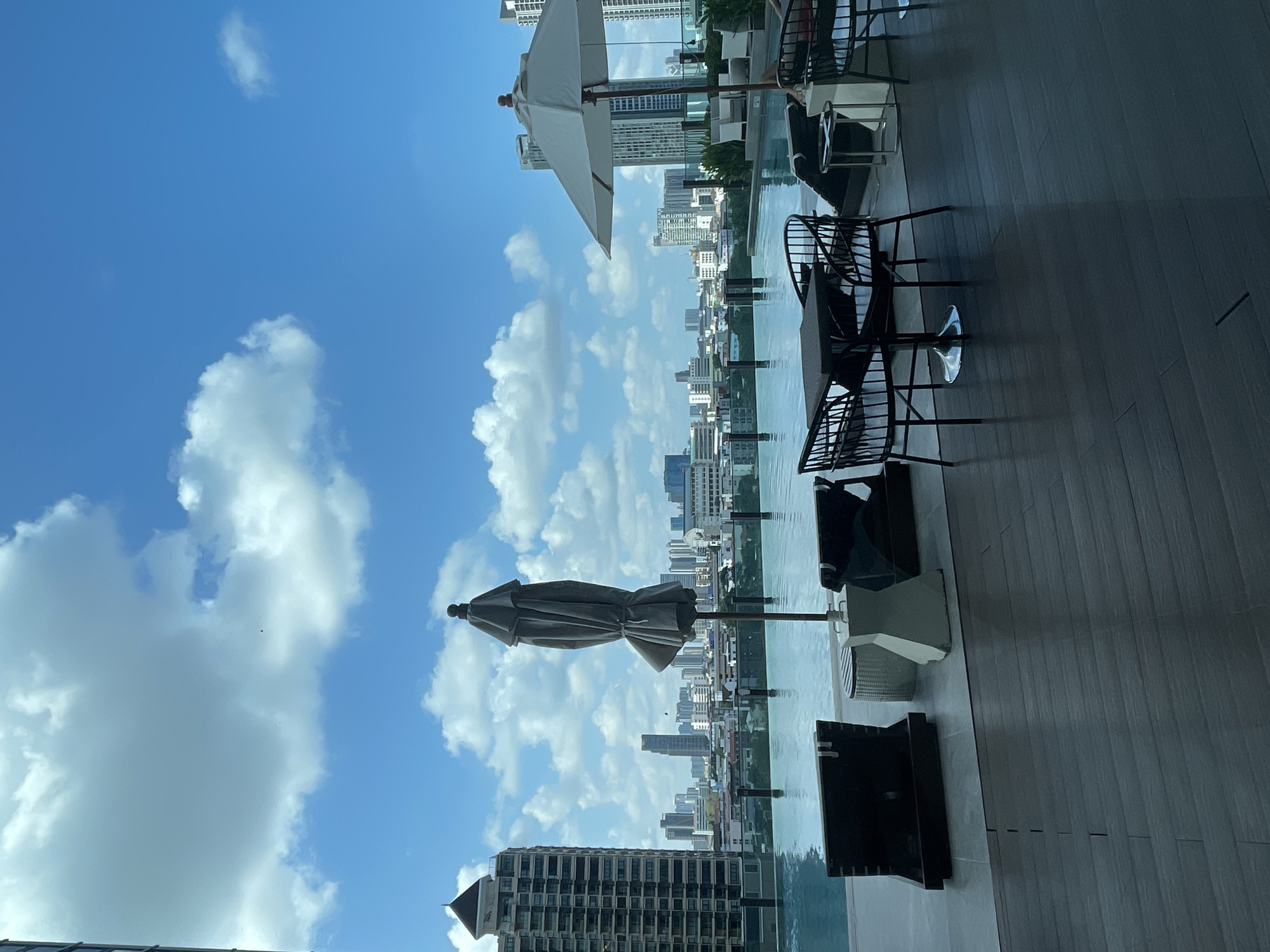 This image shows a rooftop pool area in Bangkok, Thailand, with a view of the city skyline. The scene includes outdoor furniture such as chairs and tables, as well as umbrellas. The clear blue sky contains puffy white clouds. The pool's water reflects the sky and cityscape.