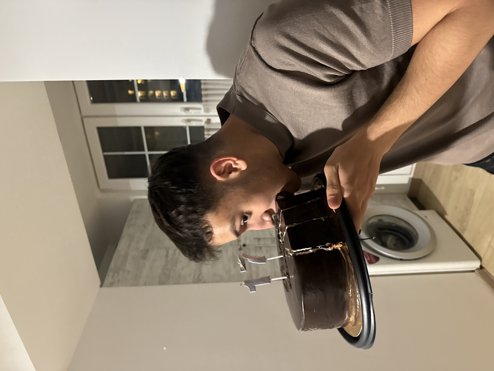 A man is holding a chocolate cake with candles in what appears to be a residential interior. A washing machine is visible in the background, suggesting a kitchen or utility area. Through a window, lights indicate an urban setting, possibly the city of Yerevan, Armenia.