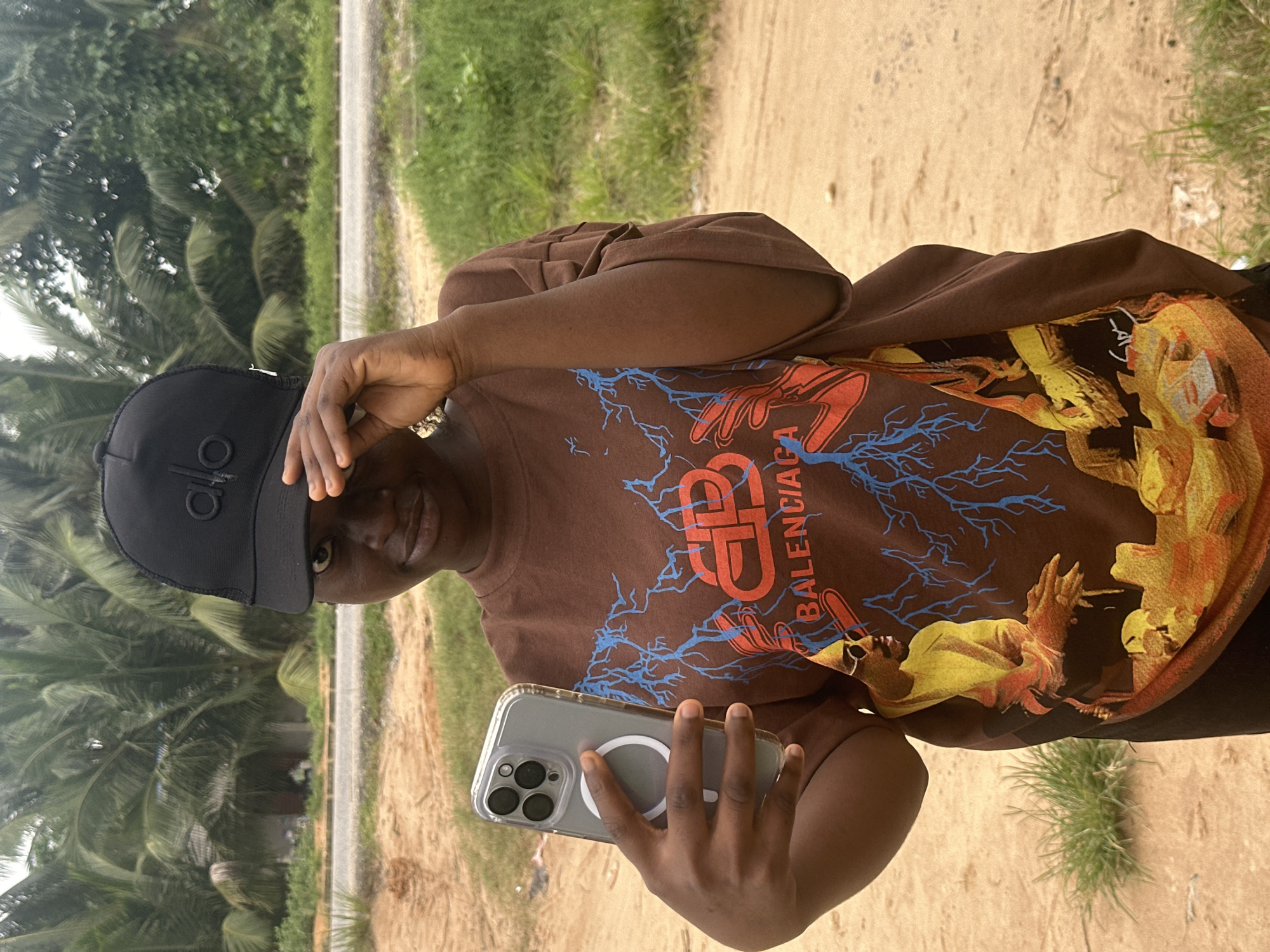 A young Black female is captured in a medium shot, standing outdoors in what appears to be a rural or semi-urban setting, possibly in Ganyingbo, Nigeria. She is wearing a brown t-shirt with a graphic print featuring abstract red and blue lightning bolts and a stylized image of a person wearing sunglasses. She also sports a black baseball cap with a logo. The woman holds a silver smartphone in her left hand, positioned as if taking a selfie, and her right hand is raised to shade her eyes from the sun. The background consists of lush green palm trees and vegetation, with a dusty path or road visible behind her. The lighting suggests it is daytime, with soft, diffused light.