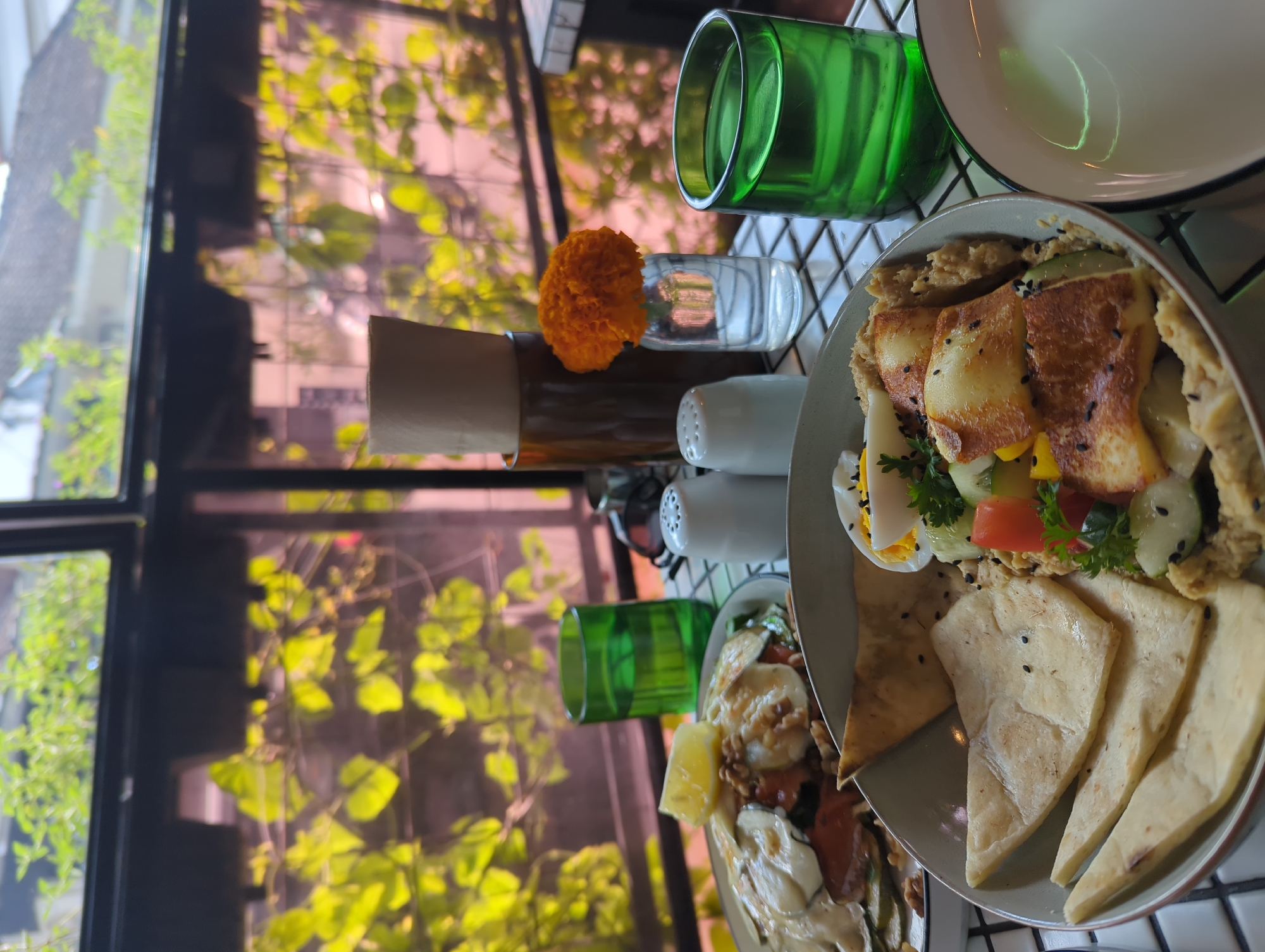 The image shows a table setting in North Kuta, Indonesia. On the table, there is a bowl of food containing pita bread, hummus, fried halloumi, sliced cucumbers, hard-boiled eggs, and diced tomatoes. Beside the plate are a green glass of water, a salt and pepper shaker set, a small vase with an orange flower, and what appears to be another dish. A window with greenery visible outside is in the background. The table itself has a black and white tiled surface.