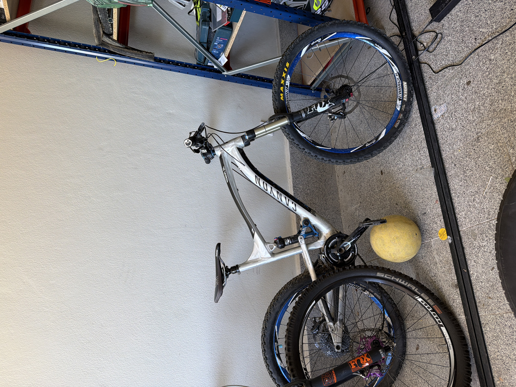 Here is a detailed description of the image:
**Scene:** The image shows a Canyon Strive AL 6.0 mountain bike in what appears to be a storage area or garage. The bike is leaning against a light gray wall to the left, with shelving visible in the upper left corner.
**Objects:**
* **Canyon Strive AL 6.0 Mountain Bike:** The bike is white with black lettering. Key features include:
* Front suspension with a FOX fork.
* Rear suspension.
* Schwalbe tires on the rear wheel and Maxxis tires on the front wheel.
* Disc brakes.
* Sun Ringle wheels.
* **Yellow ball:** Appears to be a small yellow ball, possibly a football or soccer ball, next to the bike's frame and pedals.
* **Shelving:** Located in the upper left of the frame.
* **Black metal frame:** A black metal frame is along the right side of the image.
* **Power Cord:** There is a black power cord in the top right of the frame.
**Environment:**
* **Walls:** Light gray.
* **Flooring:** Textured gray and brown, likely concrete or tile.
**Location:** The location is not specified in the image.