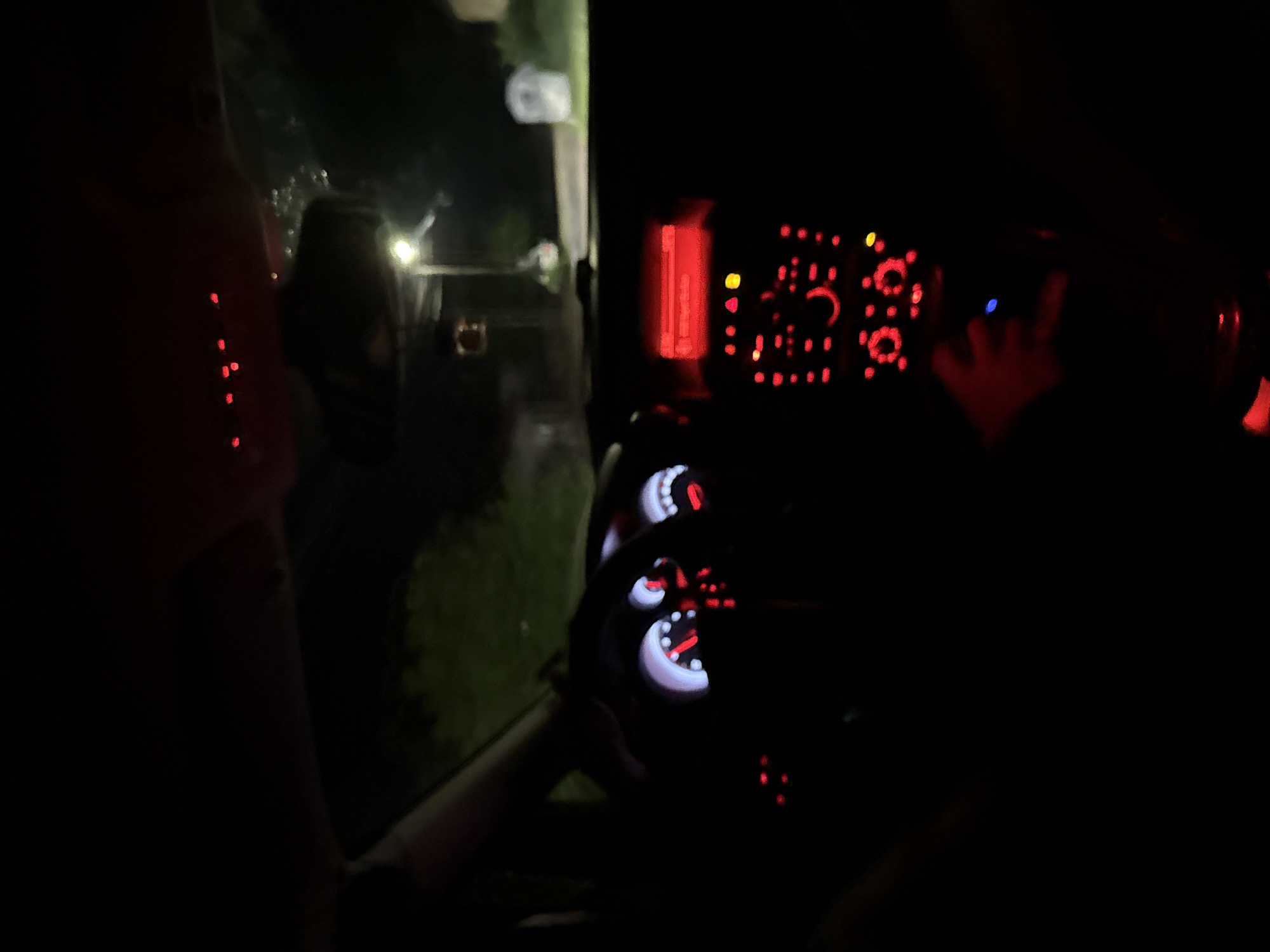 The image depicts the interior of a vehicle at night, likely in Slavyani, Bulgaria. Illuminated dashboard gauges and controls are visible, with a driver's hand near the center. Outside the vehicle, a street scene is partially visible, with a neighboring house visible.