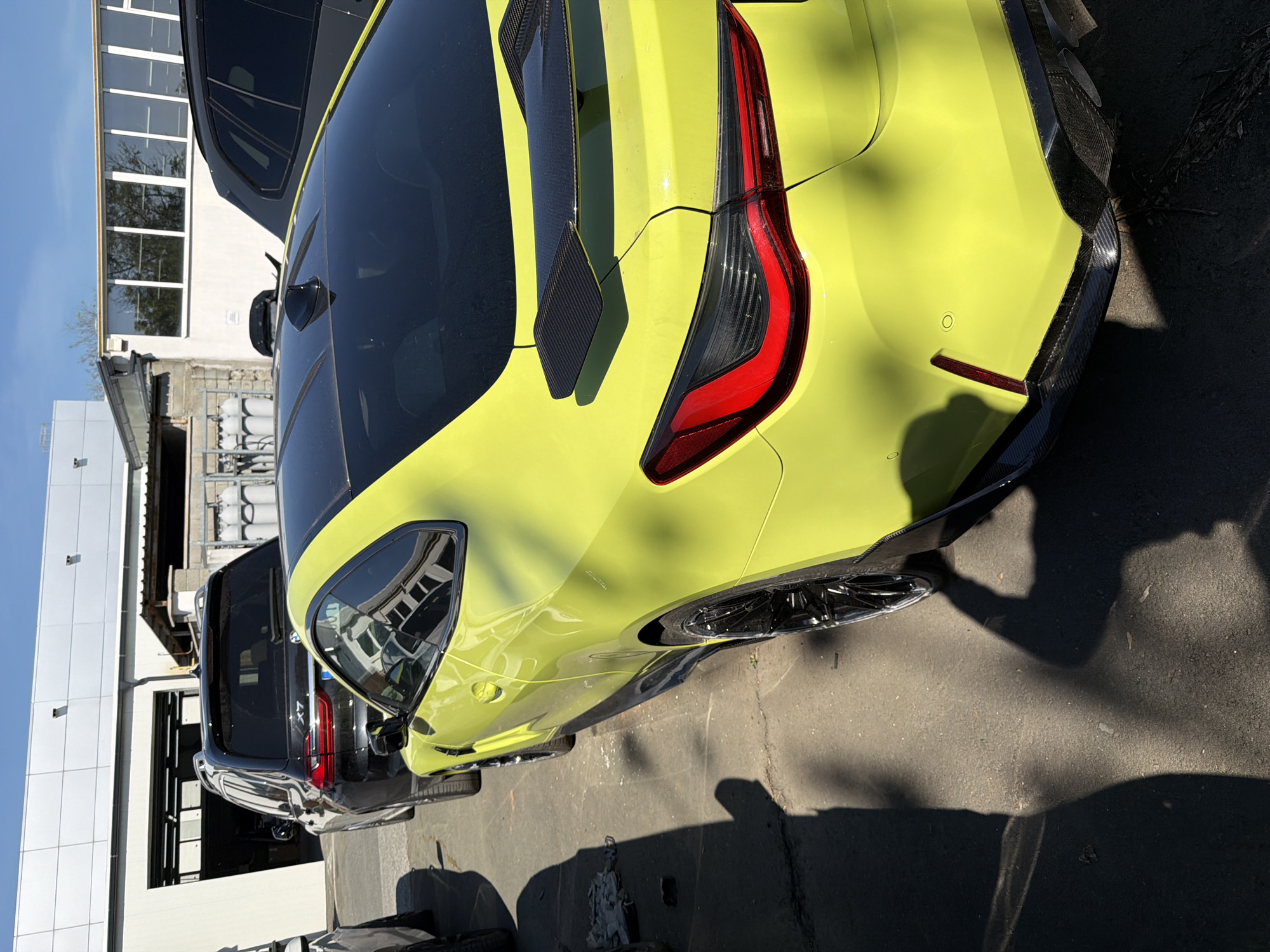 The image shows a light green BMW parked outside. A black SUV is parked behind it. The location is suggested to be in Burgas, Bulgaria. The lighting indicates it's likely daytime. There's a white building in the background with large windows. The angle of the photo suggests it was taken from a standing position close to the car. A shadow of a person is visible on the ground.