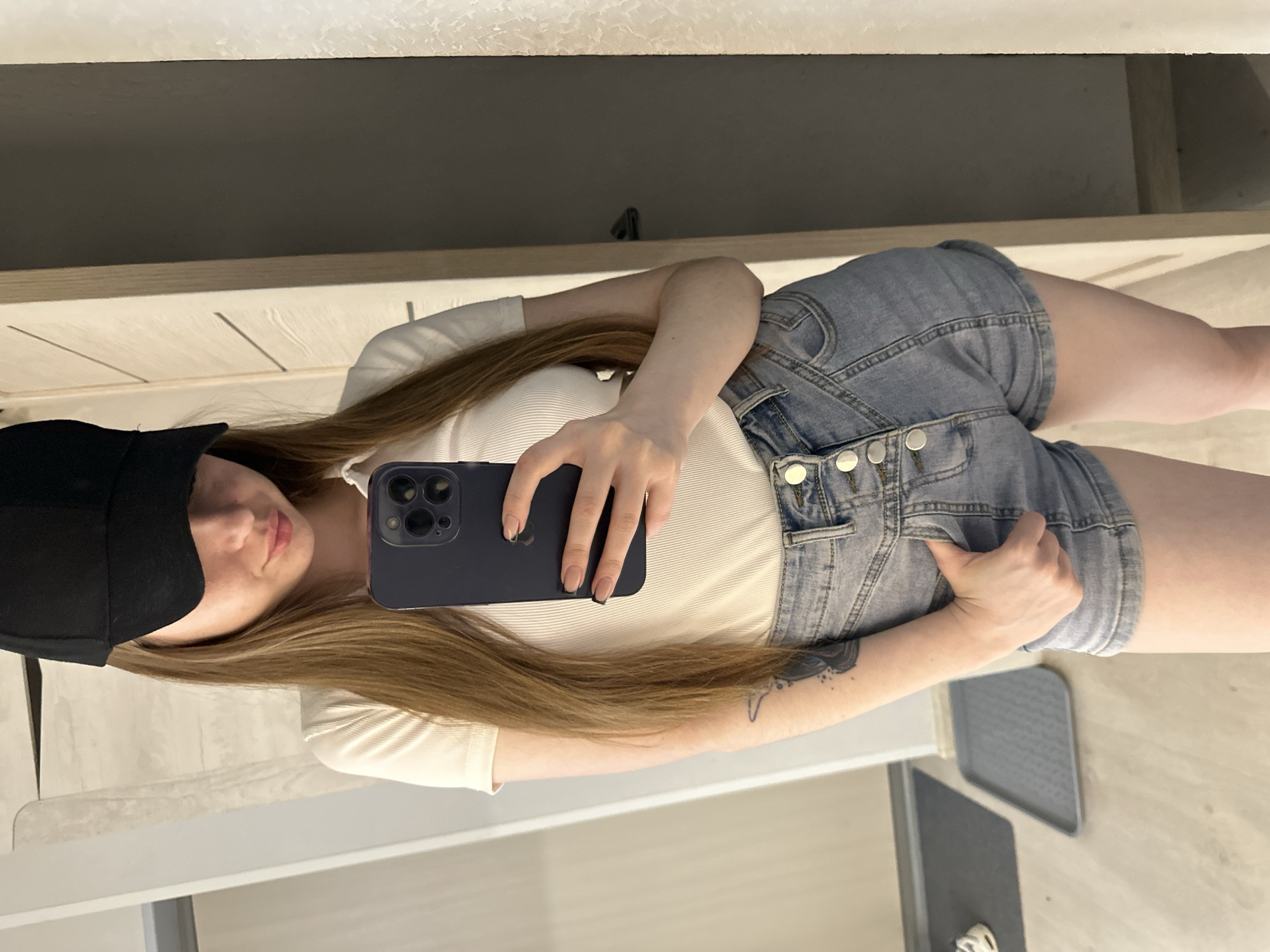 A person wearing a black baseball cap, a white ribbed shirt, and blue denim shorts is taking a selfie in an indoor environment. The person is holding a dark blue smartphone. The background includes a light-colored countertop, a darker surface above it, and a portion of a gray object on the floor.