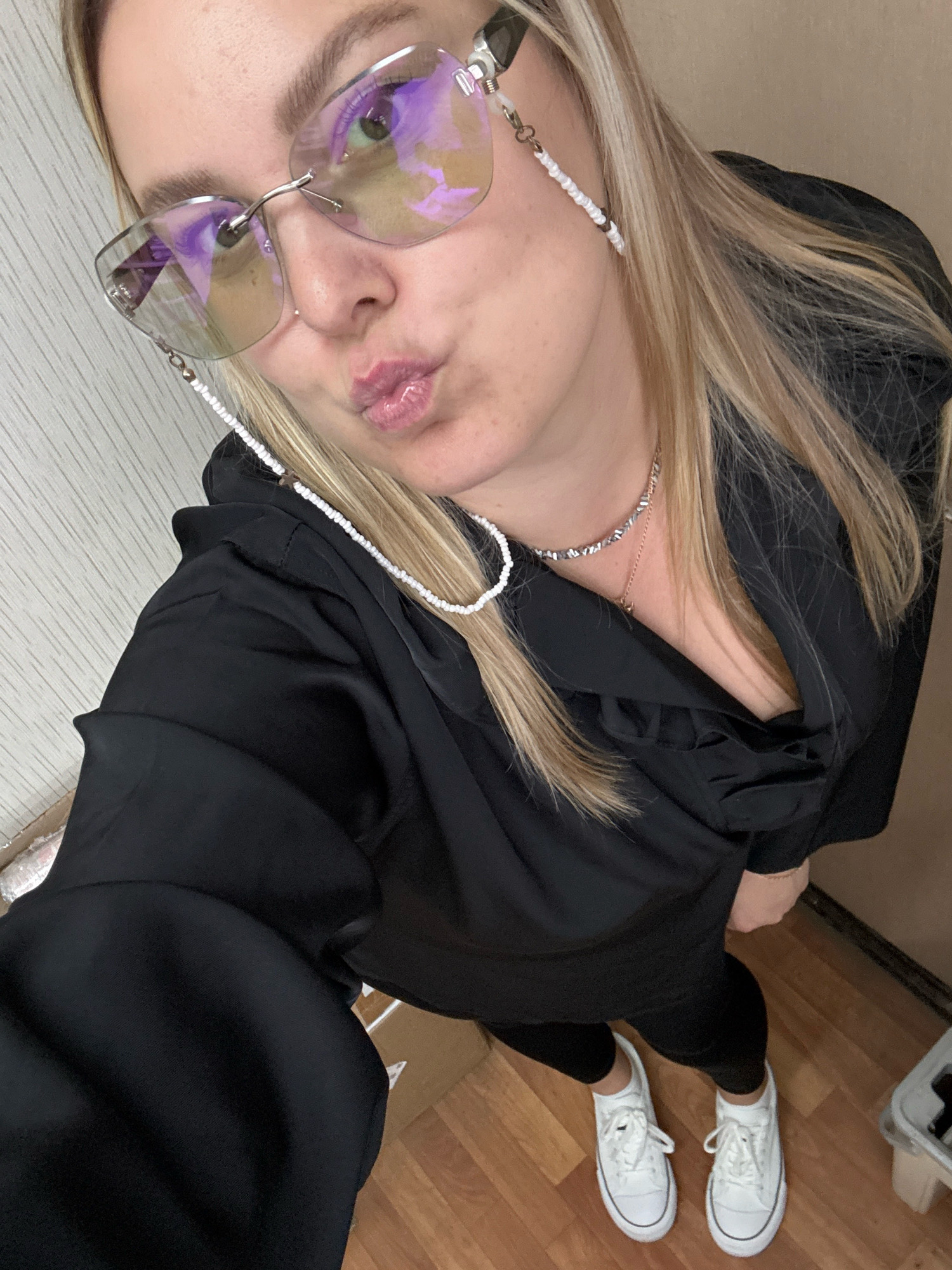 A woman with blonde hair, wearing glasses with a beaded chain, a black top with a ruffled neckline, black pants, and white sneakers, poses for a selfie. The background shows a light-colored textured wall and a wooden floor. There are also boxes visible in the lower portion of the image, one of which appears to contain packaging materials.