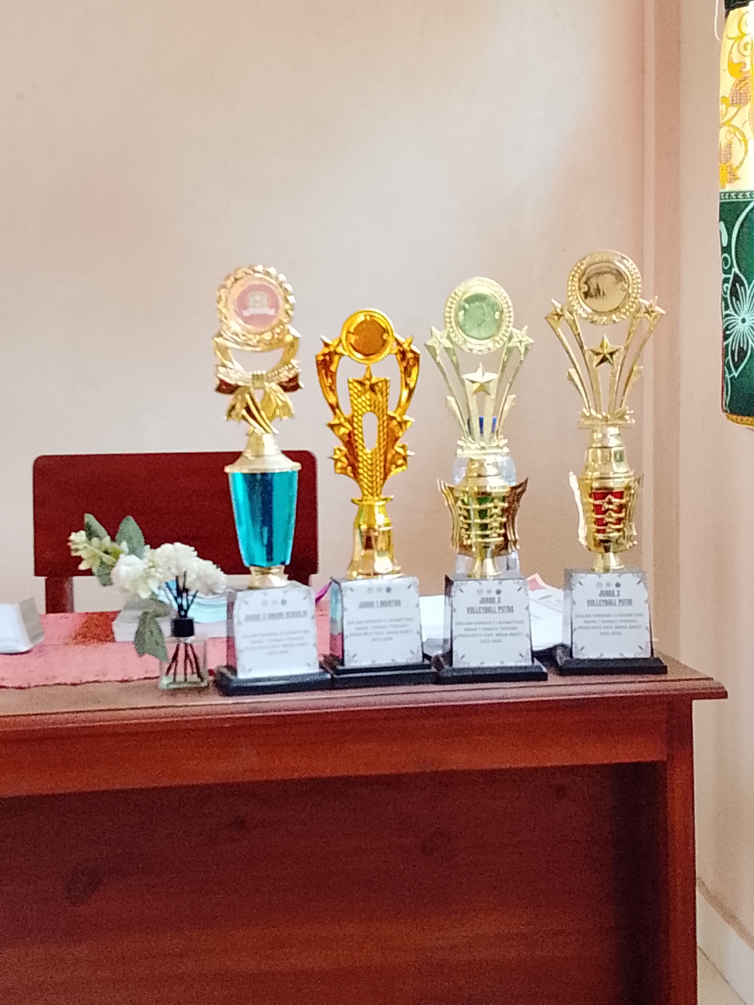Four gold and silver trophies are displayed on a wooden desk. From left to right, the trophies are gold with a blue glass cup, gold with a gold cup, gold with a green glass cup, and gold with a gold cup. Each trophy has a silver base with text. To the left of the trophies, a small vase with white flowers sits on a pink cloth. The background is a plain wall, and a portion of a red chair is visible behind the trophies. The desk is made of dark wood. A decorative fabric hangs from the right side of the frame.