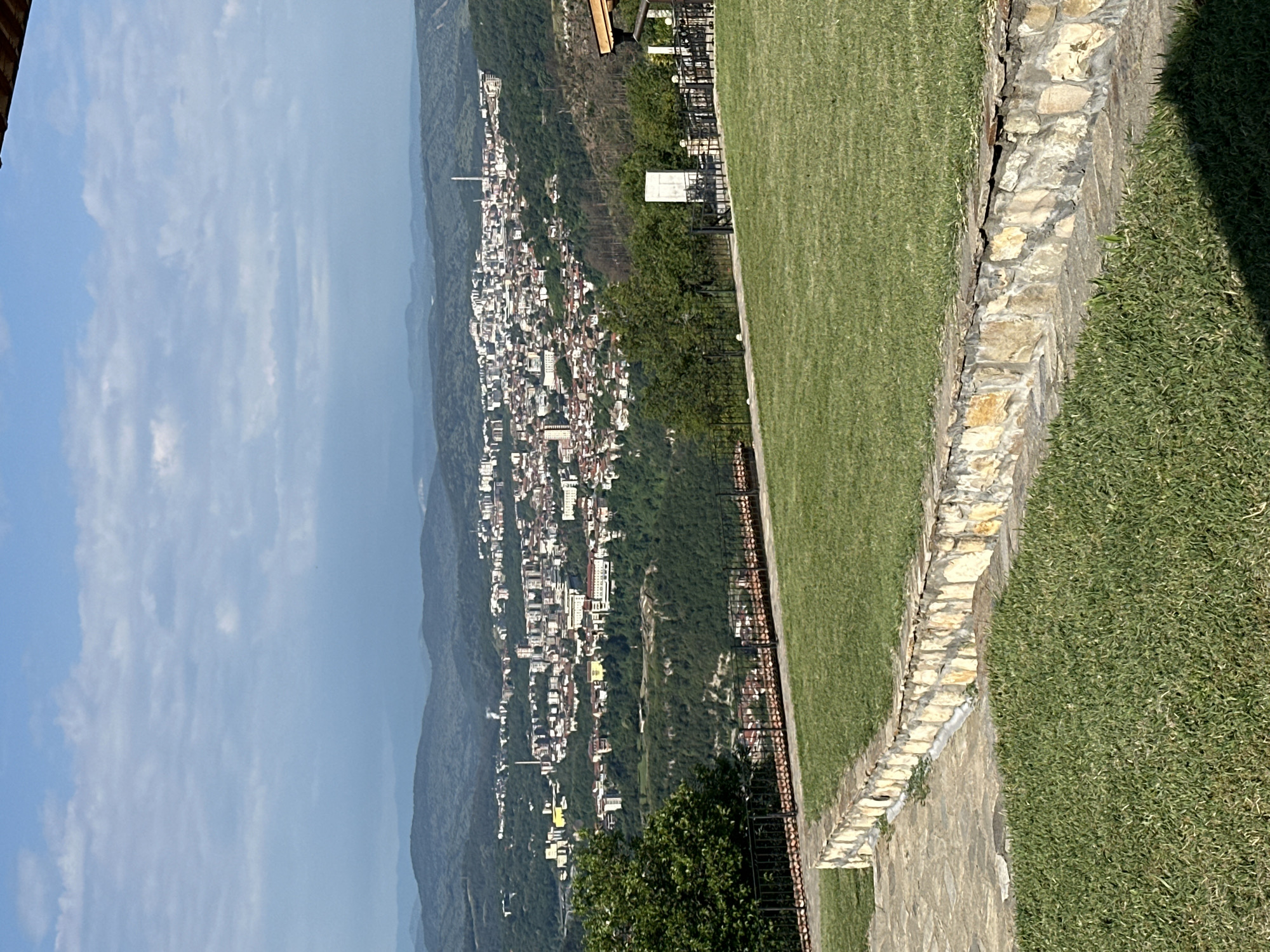 A scenic view of the city of Arbanassi, Bulgaria, is captured from an elevated position. In the foreground, a meticulously maintained lawn is bordered by a low, curved wall constructed from irregular, light-colored stones. Beyond this wall, a dark metal fence encloses another section of lawn. A mature, leafy green tree stands to the left, partially obscuring the view of the city.
The middle ground is dominated by a sprawling urban landscape, with numerous buildings of varying sizes and architectural styles nestled into the rolling hills. The city appears densely populated, with a mix of residential and commercial structures. A prominent smokestack is visible in the distance, suggesting industrial activity. The city is surrounded by lush, green forests that extend up the hillsides.
The background features a series of gently sloping, tree-covered hills that fade into a hazy, blue sky. The sky is partly cloudy, with soft, white clouds scattered across it. The overall impression is one of natural beauty and urban development coexisting harmoniously. No people or animals are visible in the image.