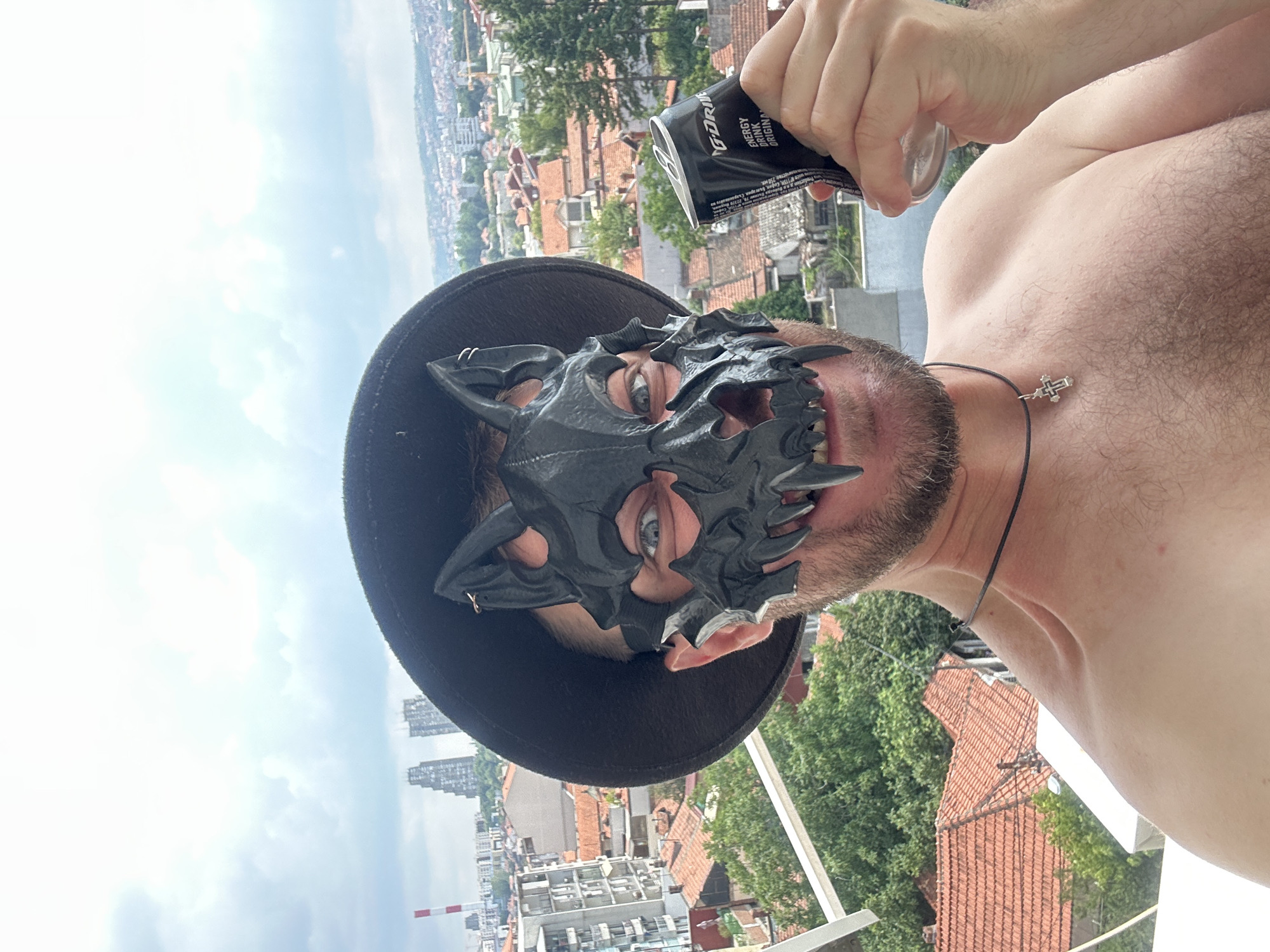 A person wearing a black dragon mask and a black hat is holding a can of energy drink. The person has blue eyes and a beard. They are shirtless and wearing a necklace with a cross pendant. The background shows a cityscape with red-tiled roofs and green trees. The sky is cloudy. The scene appears to be outdoors on a rooftop or balcony. The can has "GORILLA ENERGY DRINK ORIGINAL" printed on it. The photo seems to be taken during the daytime, likely in the late morning or afternoon. The overall mood is somewhat mysterious and playful due to the mask.