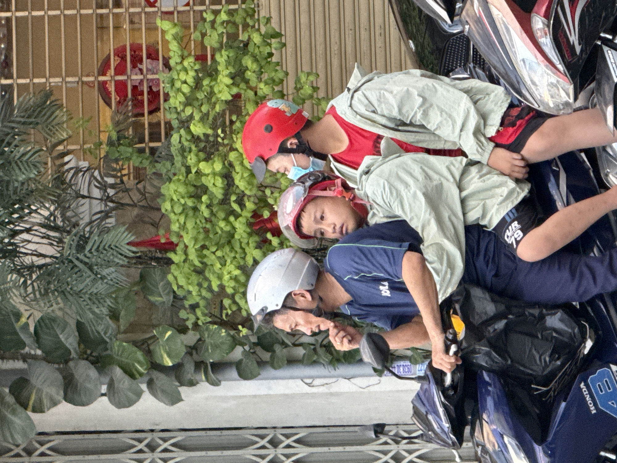 Three individuals, two boys and a man, are seated on a blue motorcycle in what appears to be a city street in Nha Trang, Vietnam. The man in the front wears a blue t-shirt and a silver helmet, his right hand resting on his chin. He is looking to his left. The two boys are seated behind him. The boy in the middle wears a light-colored jacket and a red helmet, looking forward. The boy on the right wears a red shirt and a red helmet with a cartoon character on it, as well as a light-colored jacket and a light blue face mask. He is looking to his left. The motorcycle has "HONDA" printed on its side. In the background, there are green plants, a metal gate, and a store with a closed metallic shutter. A red circular decoration is visible behind the grate.