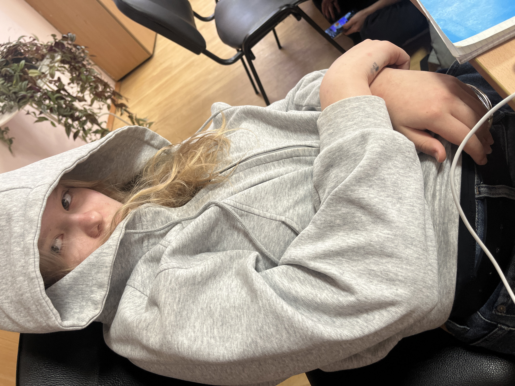 A young person is seated in a black chair, wearing a gray hooded sweatshirt and denim jeans. They are facing the camera with their hands crossed over their torso. A plant is visible in the upper left corner. Other people, a chair, a desk, a tablet, and a notebook are in the background. The environment appears to be an interior room with wood flooring. There is no evidence to suggest the location is Saint Petersburg, Russia.