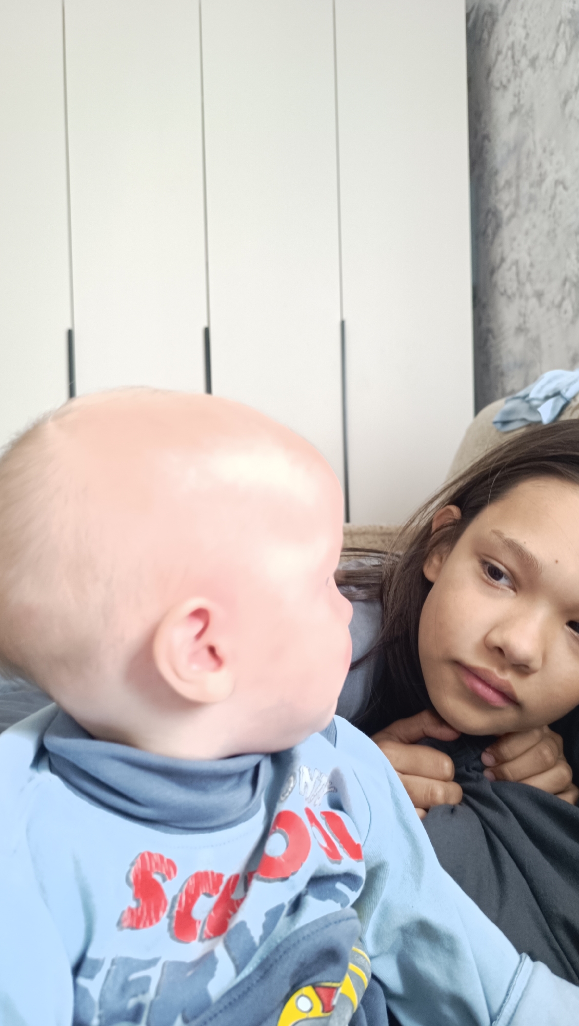 In the city of Karsha, Russia, within a home, a young child with fair skin and sparse blonde hair is positioned in the foreground, facing left, with their head turned to the right. The child is wearing a light blue long-sleeved shirt with red lettering that appears to spell "SCOTT." To the right of the child, a teenage girl with dark hair and brown eyes looks downwards, her gaze directed towards the child. She is resting her chin on her hands. The background consists of a light-colored closet with vertical lines and a textured wall. The scene appears to be indoors, likely in a living room or bedroom, with soft lighting.