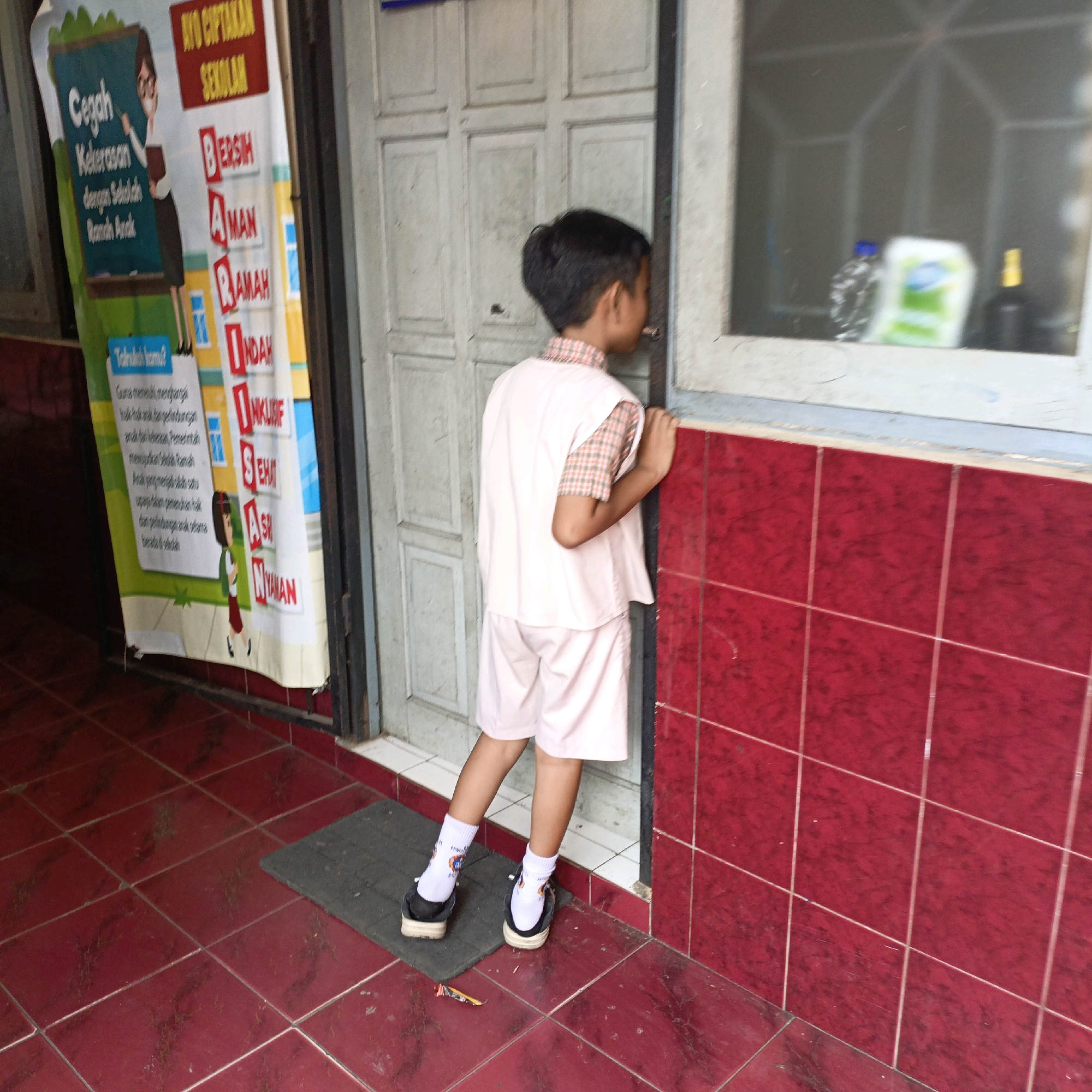 In Salatiga, Indonesia, a young boy in a light pink uniform, consisting of a short-sleeved checkered shirt and shorts, stands with his back to the camera. He is looking into a window. He wears white socks with blue and orange graphics and dark-colored shoes with white soles. A mat is placed on the red tiled floor in front of him. To his left, a banner hangs from the wall, featuring text in Indonesian and an illustration of a woman teaching children. The banner promotes "Cegah Kekerasan dengan Sekolah Ramah Anak" (Prevent Violence with a Child-Friendly School). Behind the boy is a closed door with a grid pattern. To the right of the door is a window with a red tiled wall to its right. Inside the window, obscured by the glass, are a plastic bottle, a green and white package, and a dark bottle. The floor is tiled in a repeating red pattern.