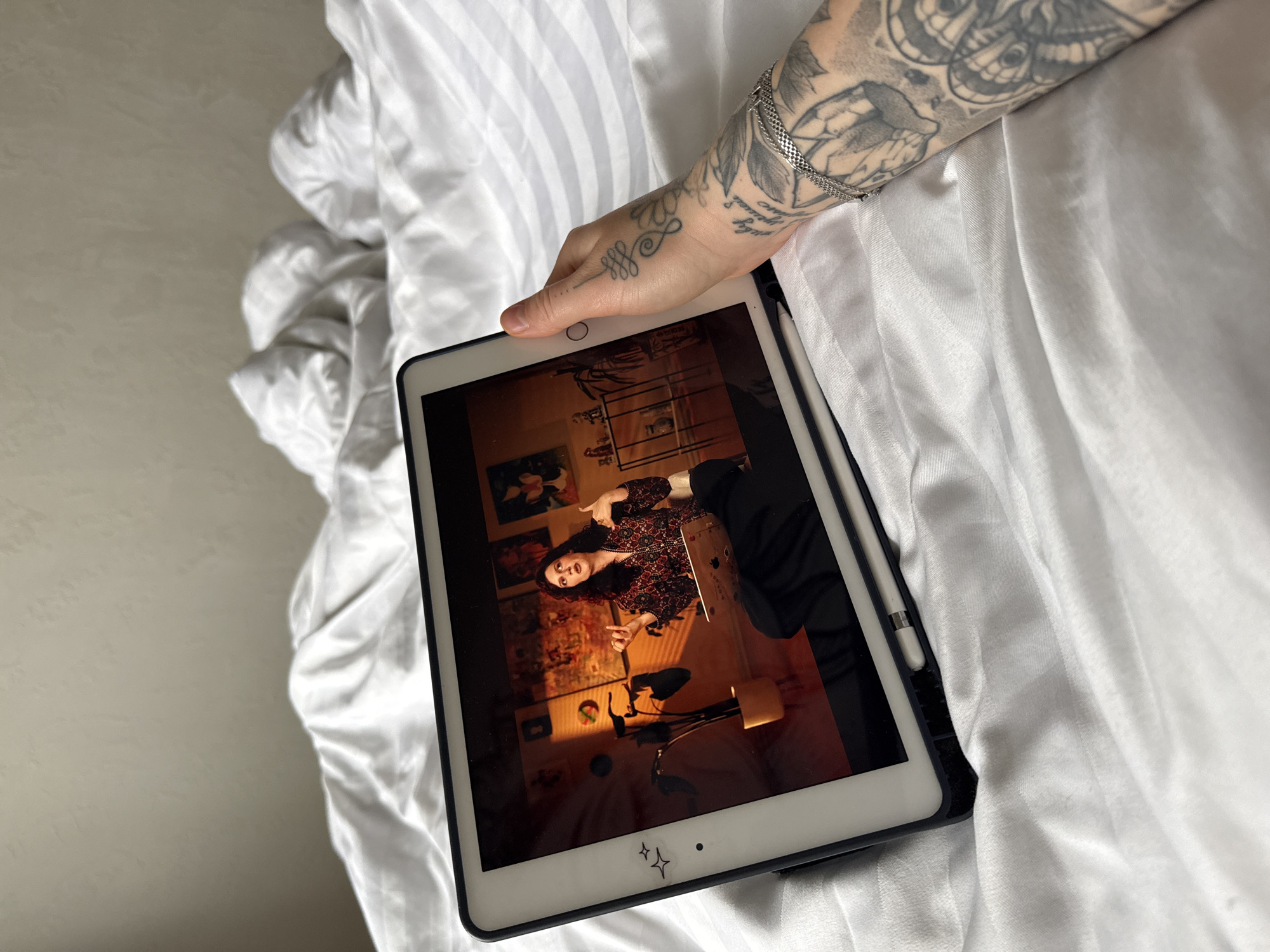 The scene depicts a personal viewing experience within a bedroom located in Rostov-on-Don, Russia. A person's left arm and hand are visible in the foreground, supporting an Apple iPad, which is encased in a dark protective cover and has an Apple Pencil attached to its side. The hand and forearm are extensively covered with black and grey tattoos, including an unalome symbol on the back of the hand and text in Cyrillic, floral, and possibly moth/butterfly designs on the arm. A silver chain bracelet is worn on the wrist.
The iPad's screen displays a video frame featuring a female individual with dark, curly hair, wearing a patterned, dark-colored top and a necklace. She is actively gesturing with both hands, one making a "call me" sign and the other open-palmed, suggesting she is speaking or presenting. A silver-colored laptop is positioned on her lap. Her background consists of a room with multiple framed artworks on the wall, a microphone on a stand, a potted plant, and a metal shelving unit displaying various small decorative items. The background within the video frame is illuminated by a warm, directional light source from the left.
The iPad and the person's arm are resting on white bedding, characterized by subtle horizontal stripes and visible folds. The background of the immediate physical environment shows a plain, light-colored wall.