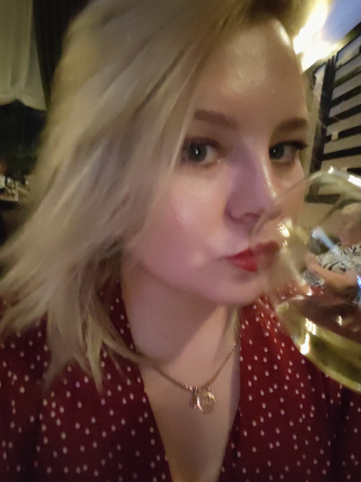 A close-up portrait captures a young woman in an indoor setting, likely a restaurant or bar in Izhevsk, Russia, during the evening. She has blonde hair and fair skin, wearing red lipstick and a dark red garment with white polka dots. A gold-colored necklace with a circular, illegible pendant is visible around her neck. Her light-colored eyes are directed towards the viewer. She is holding a wine glass containing a pale liquid, with her lips pursed as if taking a sip. In the blurred reflection on the glass, another person, possibly an older woman, wearing a patterned top, can be faintly seen in the background. The lighting is dim, suggesting a relaxed social atmosphere.