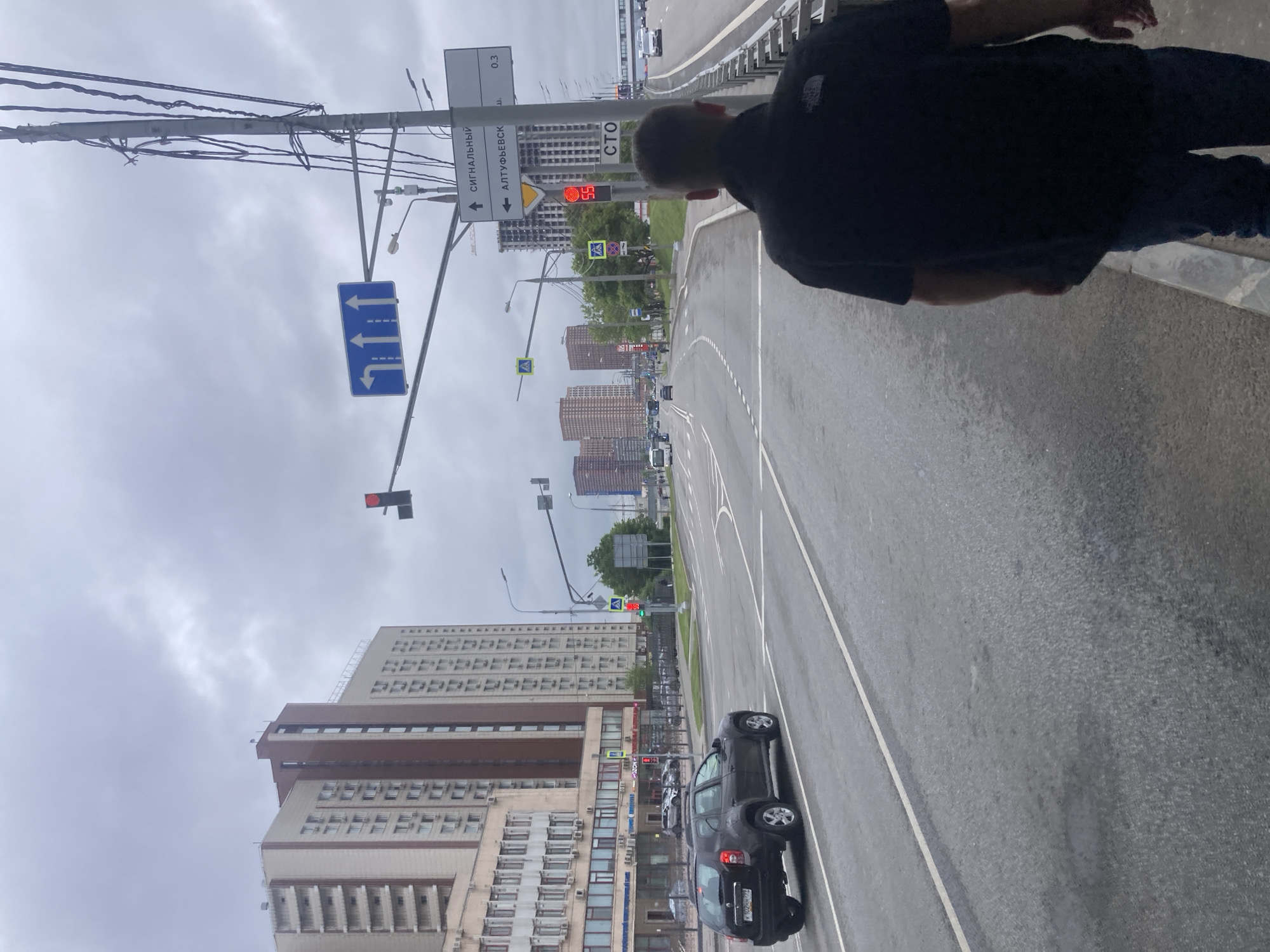 The image shows a street scene in Khimki, Russia, under an overcast sky. A man wearing a dark-colored shirt and jeans is standing on a grey concrete platform, partially obscuring the view. The street features a multi-lane road with white lane markings. Traffic includes at least one dark-colored SUV with visible license plate details. On the left side of the road, there is a large building with a light-colored facade and multiple rows of windows. Street signs are visible, including one indicating directions and another pointing towards Altufyevsk highway. There are traffic lights above the road, one showing a red signal. Further down the road, several high-rise buildings are visible. Power lines stretch across the sky.