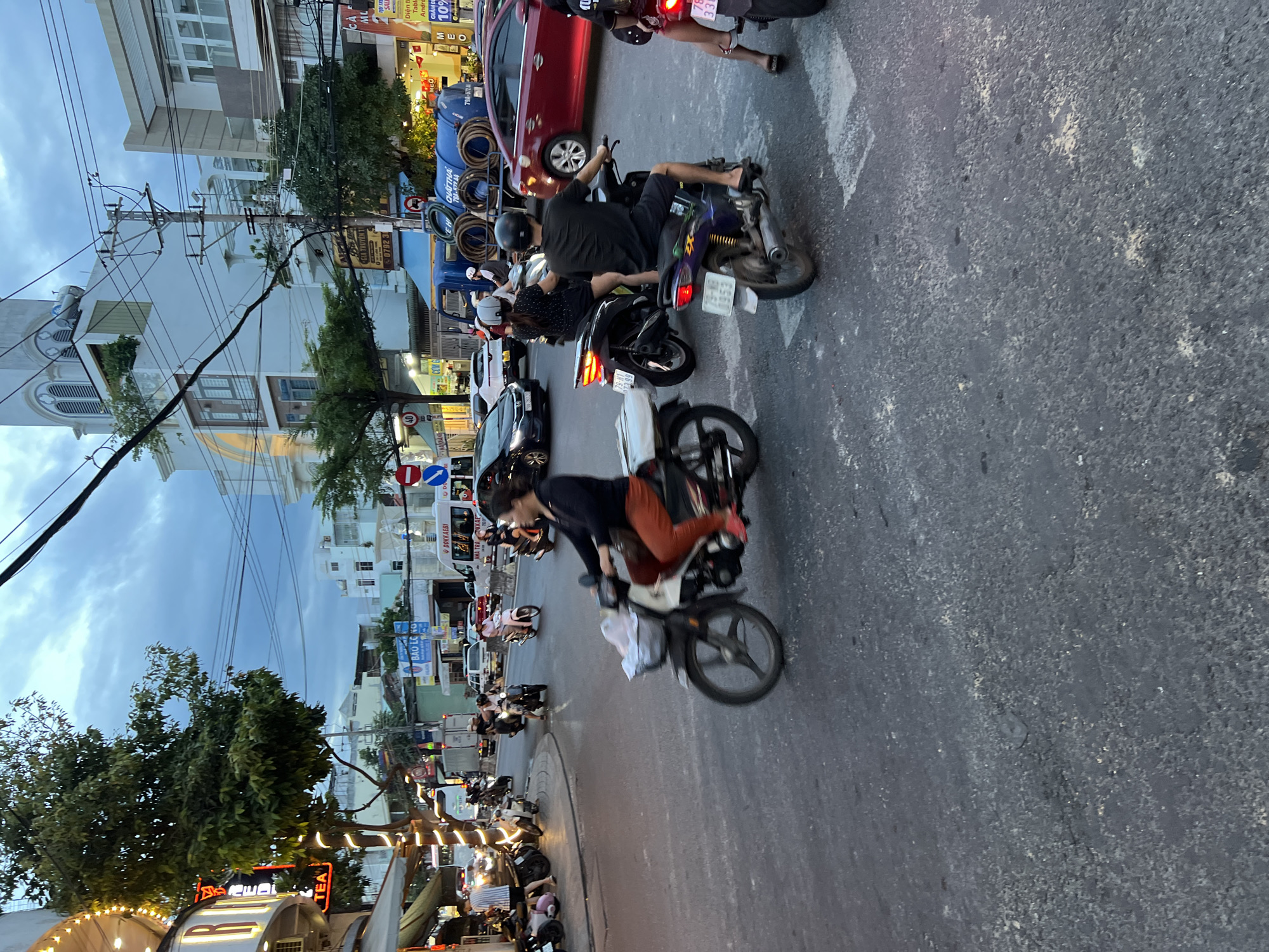 This is a street-level, eye-level, medium-shot, outdoor photograph taken at dusk in Nha Trang, Vietnam. The image captures a busy intersection with a blur of motion from multiple motorcycles and cars. In the foreground, a person wearing an orange shirt and black pants is riding a white motorcycle with a basket on the front, moving from left to right. To their right, a person in a black shirt and shorts is riding a purple motorcycle. Further right and slightly behind, another person on a motorcycle is visible, and to the far right, a portion of a person on a red motorcycle is seen. In the background, numerous other motorcycles, cars, and a large blue truck with hoses are navigating the intersection. Buildings line the street on either side, some with illuminated signs and awnings, indicating commercial activity. The sky is overcast with a dusky hue. The scene suggests a typical evening commute or busy period in the city.
