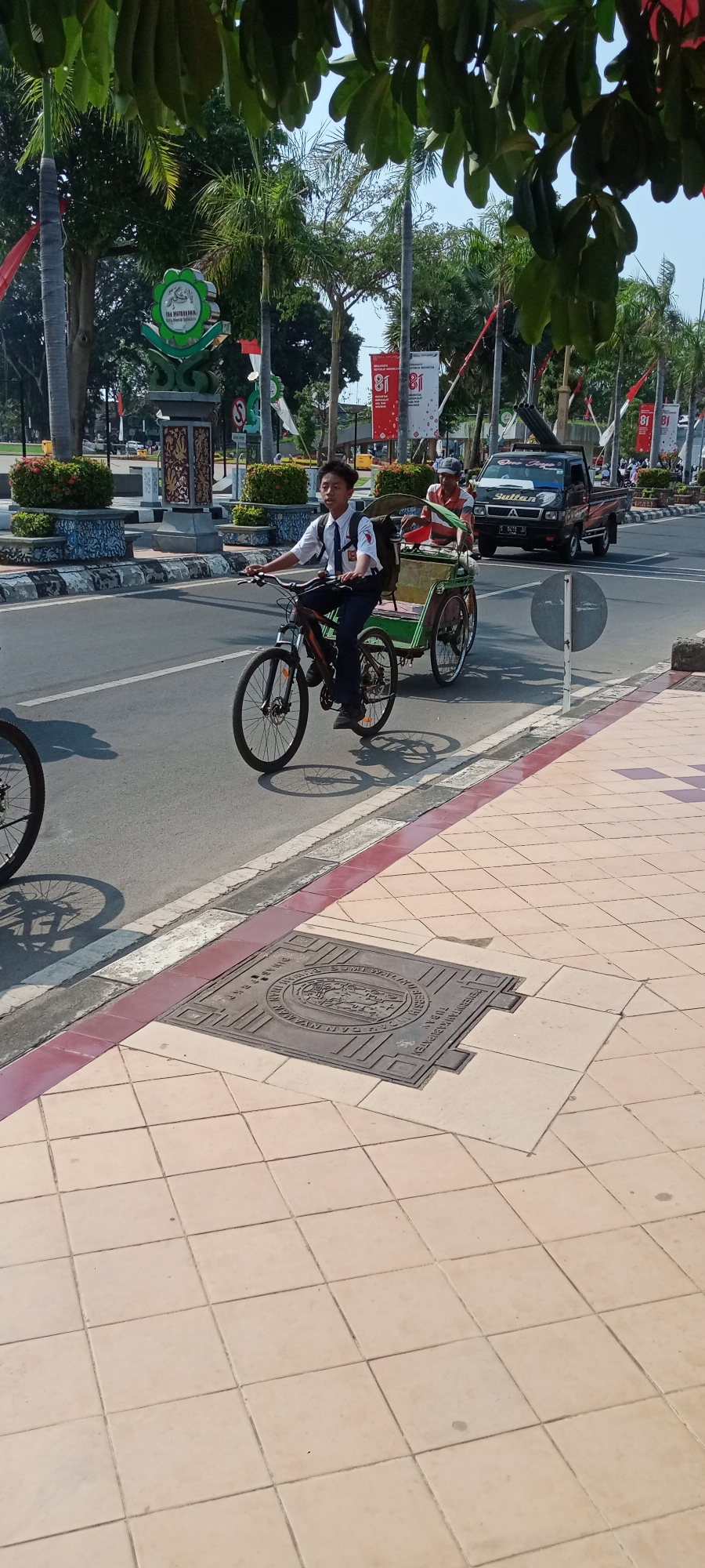 An outdoor, eye-level, medium shot captures a street scene in Tuban, Indonesia, under a clear, bright sky. In the foreground, a young male wearing a white school uniform with a dark tie and dark pants is riding a bicycle. He is pulling a rickshaw with a green canopy, in which an older male wearing a striped shirt and a cap is seated. The bicycle and rickshaw are moving on the asphalt road, casting distinct shadows on the pavement. The pavement is tiled with square, light-colored tiles, with a red border running along the edge of the road. A manhole cover with embossed text and a circular emblem is visible on the pavement. In the background, a black pickup truck with the name "Sultan" on its side is driving on the road. Several palm trees and other greenery line the street, along with decorative bushes. Banners with the number "81" in red and white are displayed on poles along the sidewalk. A monument with a green circular emblem is visible to the left. The overall atmosphere suggests a sunny day with moderate activity on a city street.