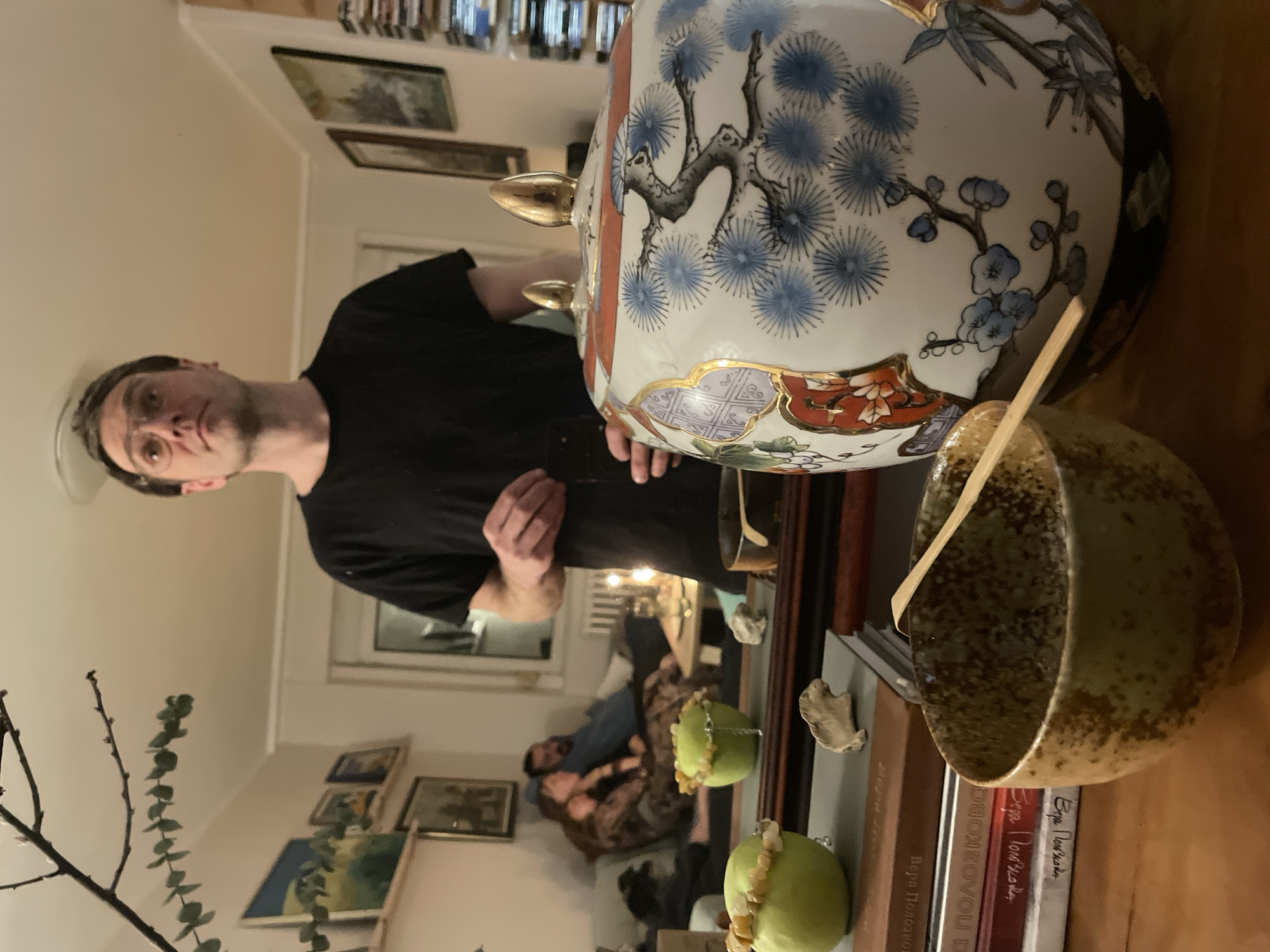 The scene is an indoor setting, possibly a living room in Belgrade, Serbia. The foreground features a large, ornate vase with floral and geometrical patterns. Next to it is a ceramic bowl with a wooden spoon inside. The bowl is placed on top of several books. Behind the vase and bowl, a man wearing a black t-shirt and a hat is visible. He appears to be looking at the camera. In the background, reflected in what appears to be a mirror, are two people relaxing on a couch. The surrounding environment includes framed pictures on the wall, a branch with leaves, and a bookshelf filled with books.