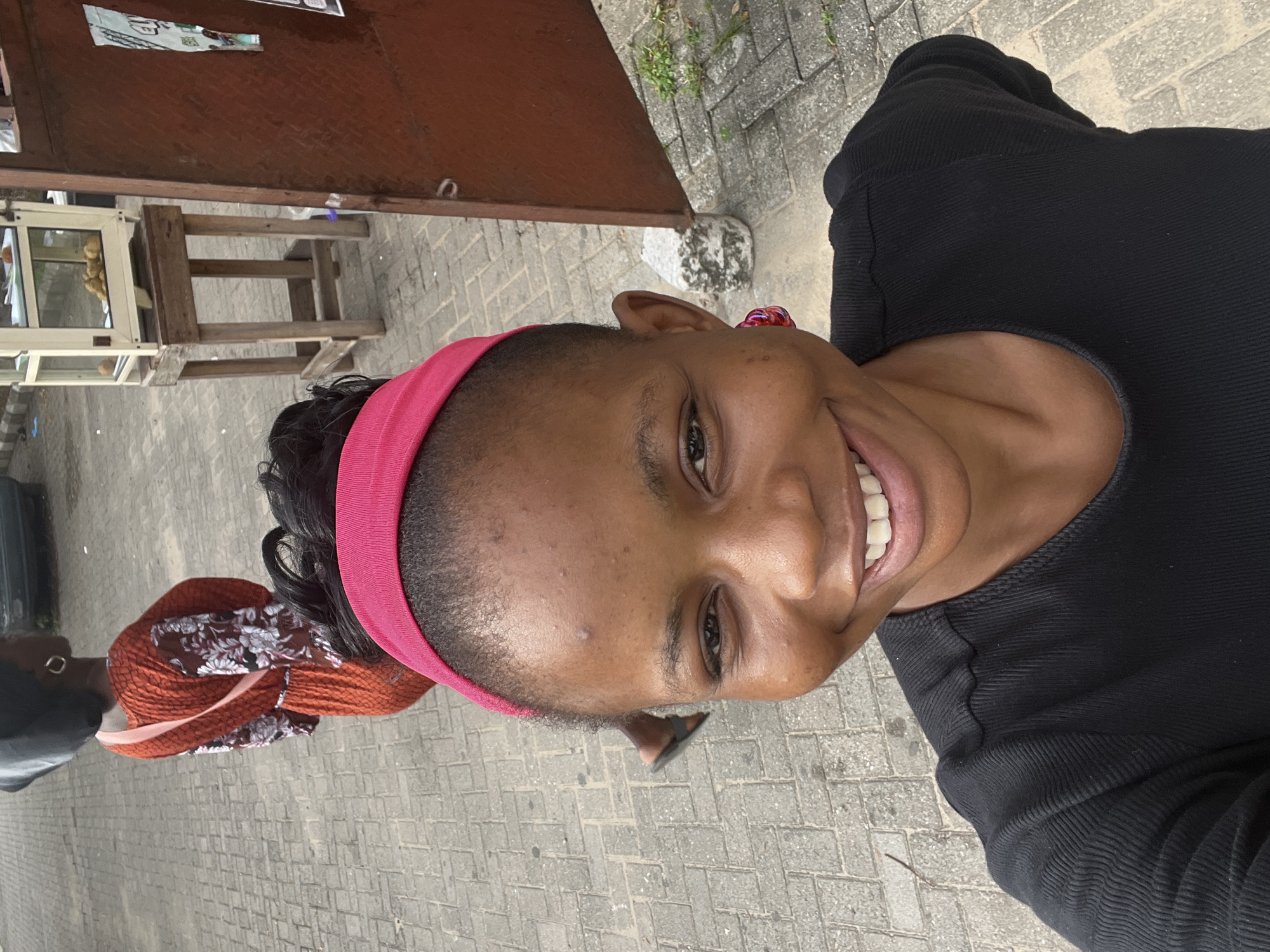 A medium shot shows a smiling, dark-skinned woman wearing a pink headband and black top. She is looking directly at the camera. The scene is outdoors on a paved area. Behind her is a metallic red structure and a wooden table with food on display. Another person with a brown head covering and black shirt is walking behind her. The image appears to be taken in Ijanikin, Nigeria.