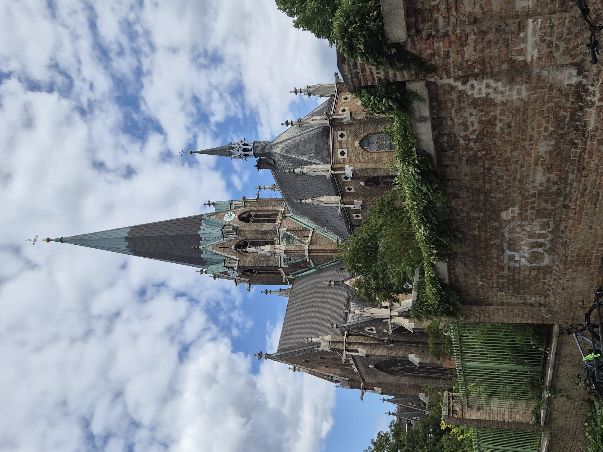 The image depicts St. Mary's Assumption Church (Mariä-Himmelfahrt-Kirche) in Vienna, Austria. The church is a prominent Gothic Revival structure with a tall, dark spire topped with a golden cross and globe. The building is constructed of brick and features intricate architectural details, including pointed arches, gargoyles, and smaller spires. The sky is partly cloudy with white cumulus clouds against a blue background. In the foreground, a low, green metal fence runs along a weathered brick wall. There are lush green trees and foliage bordering the church and growing over the wall. Two bicycles are parked in front of the wall on the lower left of the image. The overall scene captures a sunny day in front of an impressive religious building.