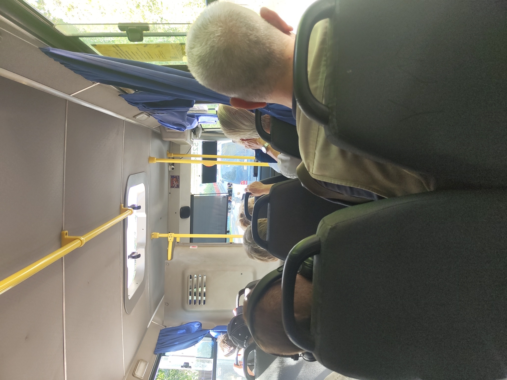 The image depicts the interior of a bus, likely in Ramenskoye, Russia, as indicated by the context of the original query. The perspective is from a passenger seat, looking forward. Several passengers are visible, their faces mostly obscured. The seats are upholstered in a dark fabric. Yellow safety poles and handrails are prominent throughout the bus. Blue curtains hang along the windows. Outside the windows, foliage and other vehicles are visible, suggesting the bus is in motion. The lighting appears to be natural daylight filtering in. There are no discernable interactions between the passengers shown. The overall scene is mundane, representing typical public transportation.