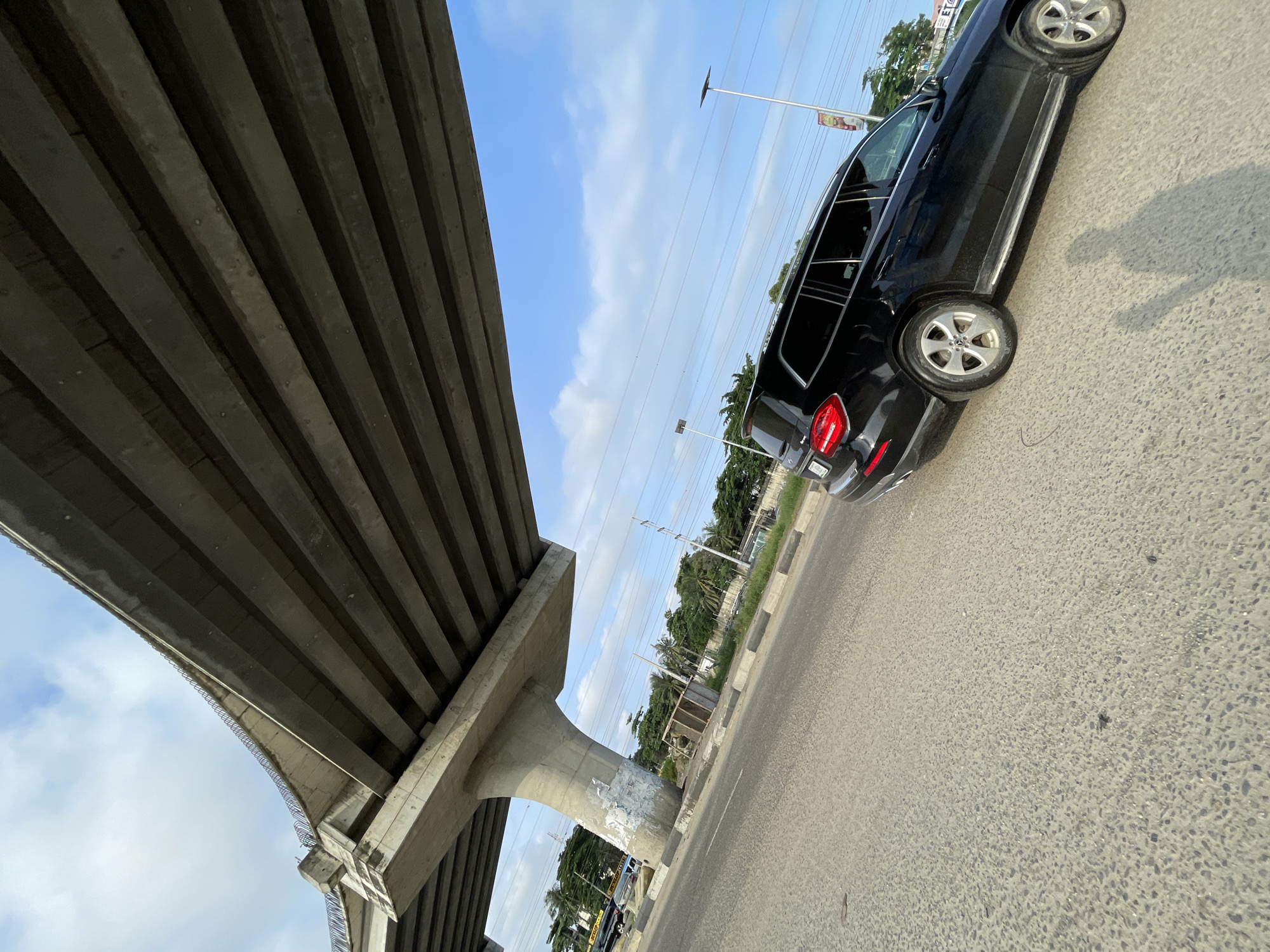 A black Mercedes-Benz GLS SUV is parked on the side of a road in Lekki, Nigeria, beneath an elevated concrete highway currently under construction. The vehicle faces right, with its rear passenger side closer to the camera. The road is paved with asphalt and has visible white and grey curbs. Tall trees and foliage line the background, with some utility poles and power lines visible. The sky is blue with scattered white clouds. The shadow of a person is cast on the road to the left of the SUV. There is some graffiti on the concrete support pillar of the highway.