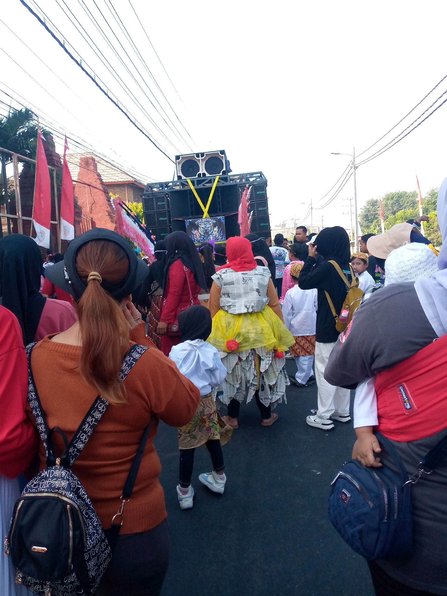 The image captures an outdoor scene in the city of Sumberwono, Indonesia, likely during a cultural event or parade. A large speaker system is set up, suggesting a public gathering or performance. Several people are present, with a focus on individuals in traditional or celebratory attire. A child in the foreground wears a unique costume made of newspaper and yellow fabric, adorned with red pom-poms. The crowd includes women in hijabs and men, some wearing traditional Indonesian clothing like batik. Red and white Indonesian flags are visible, further indicating a nationalistic or festive occasion. The environment appears to be a street, with overhead power lines and some buildings in the background. The lighting suggests it is daytime.