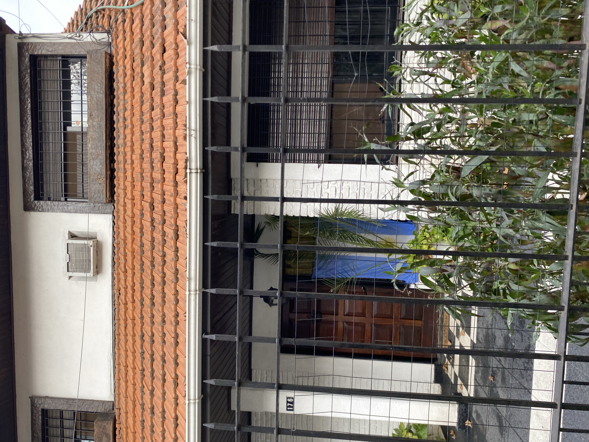 The image shows the facade of a house with an orange tile roof, located in Martínez, Argentina. A black metal fence is in the foreground, partially obscuring the view of the house. Behind the fence, there is a white brick wall, a wooden front door, and windows with dark blinds. The number "176" is visible on the white wall to the left of the door. A portion of the Argentinian flag, a blue and white striped banner, is hanging to the right of the door, near a green plant. An air conditioning unit is installed in the upper part of the white wall, above the roofline. There is a second window with dark blinds on the upper floor. The ground in front of the door has a checkered pattern.