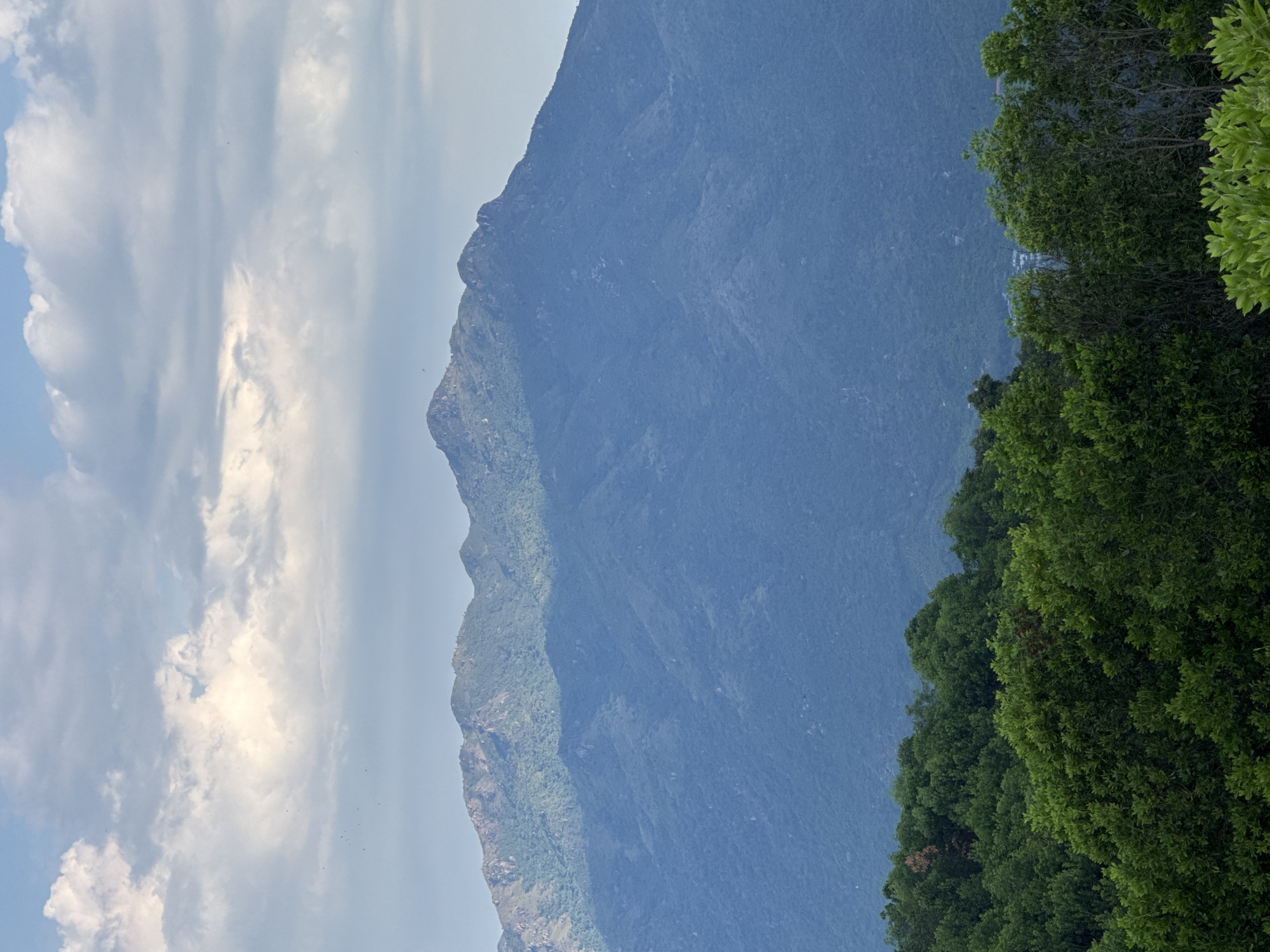 The image shows a panoramic view of a lush green forest in the foreground, with a dense canopy of trees. Beyond the forest, a majestic mountain range rises, its slopes covered in vegetation. The mountain peaks are silhouetted against a sky filled with dramatic clouds, some brightly illuminated by sunlight and others casting shadows. A portion of the mountain range appears to be in shadow, suggesting a specific time of day or weather conditions. In the distance, a small cluster of buildings can be seen, hinting at a settlement nestled within the landscape. The overall scene evokes a sense of natural beauty and vastness, characteristic of the mountainous terrain surrounding Nam Nha Trang Ward, Vietnam.