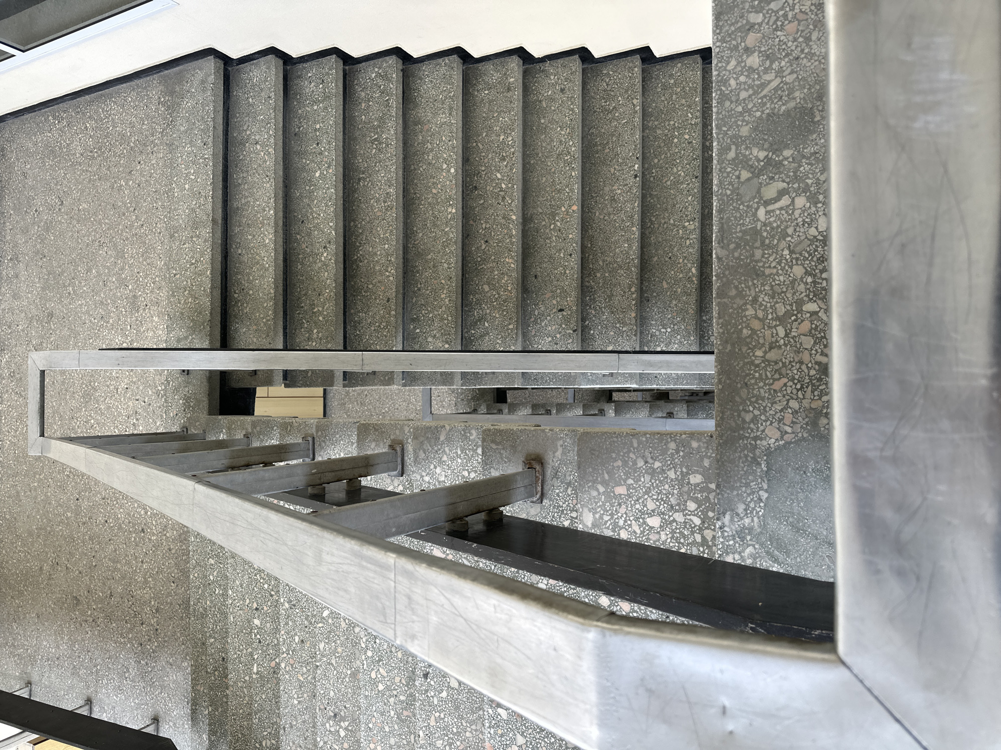 The image is a high-angle shot of a staircase, likely indoors, with a focus on the stairs and the metallic railing. The steps are made of a textured material, possibly terrazzo, in shades of gray with darker edges. The railing is constructed from metal, appearing to be stainless steel or a similar material, with horizontal supports and vertical posts. The environment surrounding the staircase is also made of the same textured material as the steps. There are no people visible in the image, and the focus is solely on the architectural elements of the staircase. The location is not evident from the image, thus, the presence in Varna, Bulgaria, cannot be confirmed from the visual content alone.