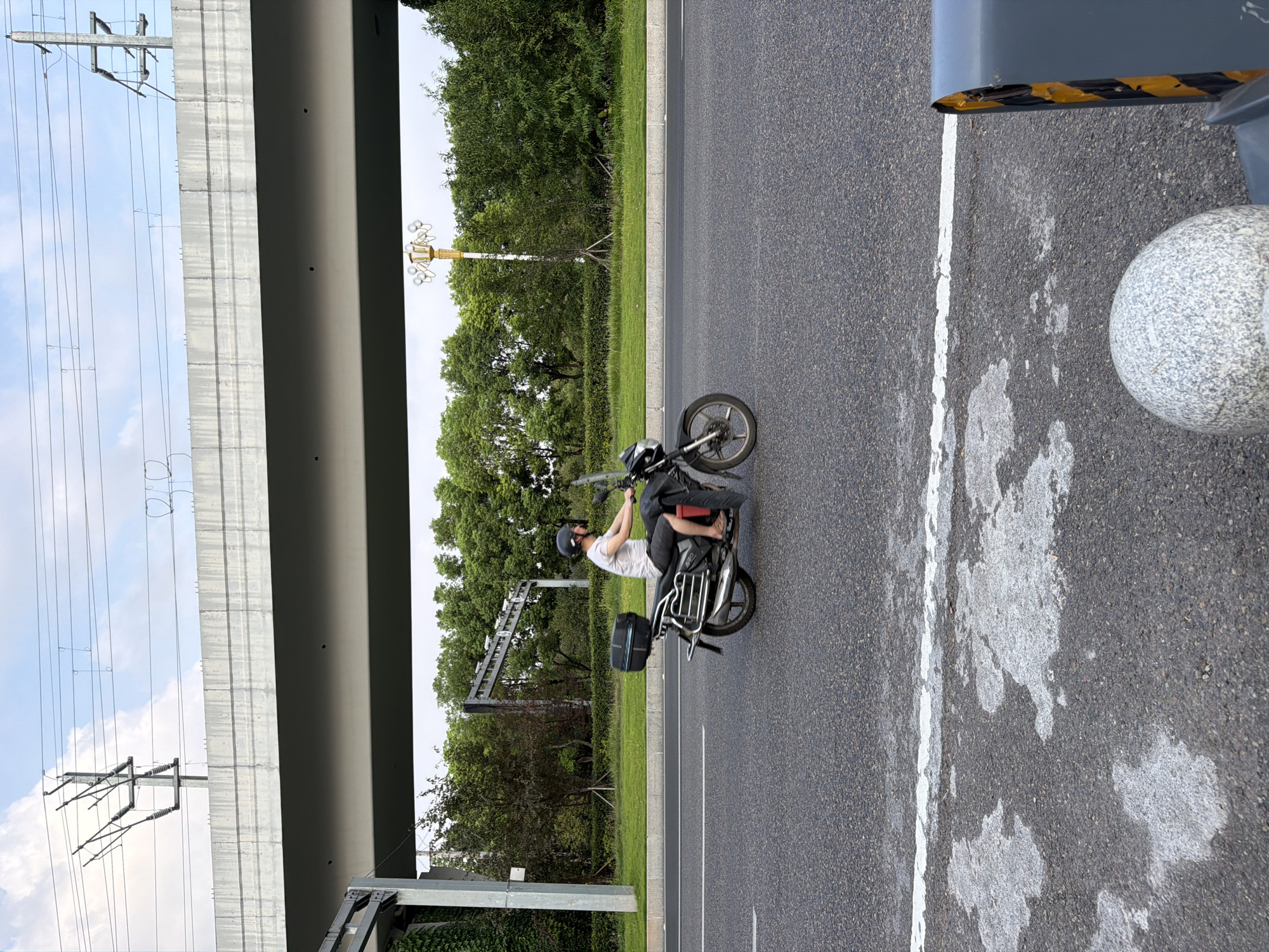 A person wearing a helmet, t-shirt, and shorts rides a black motorcycle with a black cargo box on the back. The motorcycle is traveling on a paved road with a white line. In the background, there is a large elevated concrete structure, trees, and a street lamp. The sky is visible above. This image was taken in Jiangjin, China.