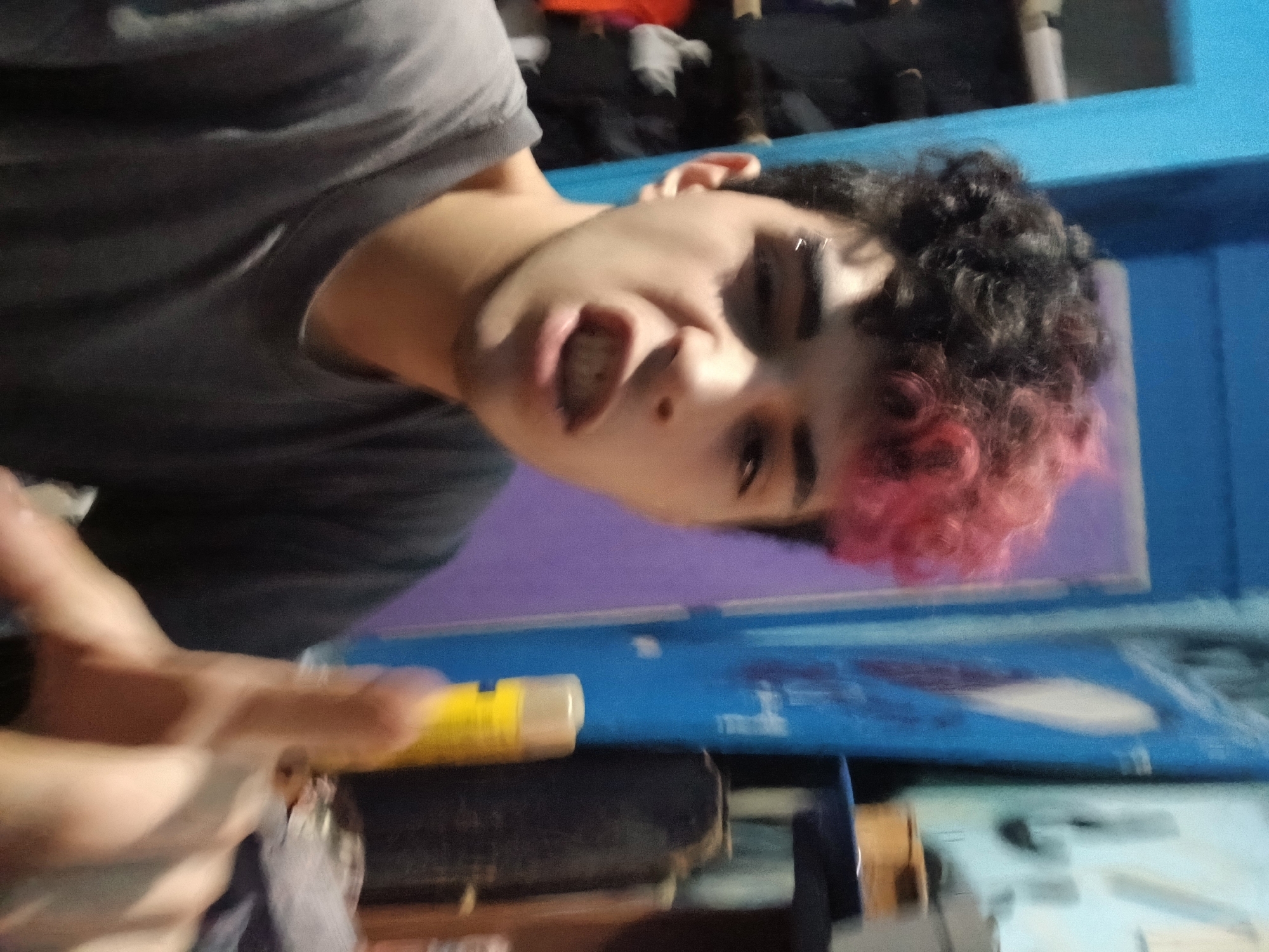 A close-up, low-angle shot depicts a person with fair skin, dark curly hair with dyed pink tips, and an eyebrow piercing on their left eyebrow. They are wearing a gray t-shirt and have their mouth slightly open. The person is holding a yellow and clear object.
The background includes a blue wall, a purple wall, and assorted items that are mostly out of focus. The location in the city of Buenos Aires, Argentina is unknown.