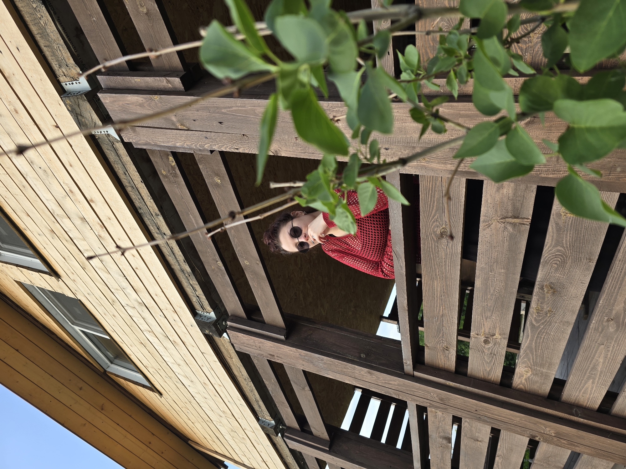 An outdoor, low-angle, medium shot captures a woman standing on a wooden deck of a wooden house. The woman is wearing a red, crochet-like sweater and dark sunglasses. She is looking to her left with her chin resting on her hand. The wooden structure is made of dark brown, weathered planks. In the foreground, a green plant with leaves partially obscures the view. The background shows the siding of a wooden house and a clear blue sky. The scene appears to be in Ермолаевский Затон, Russia.