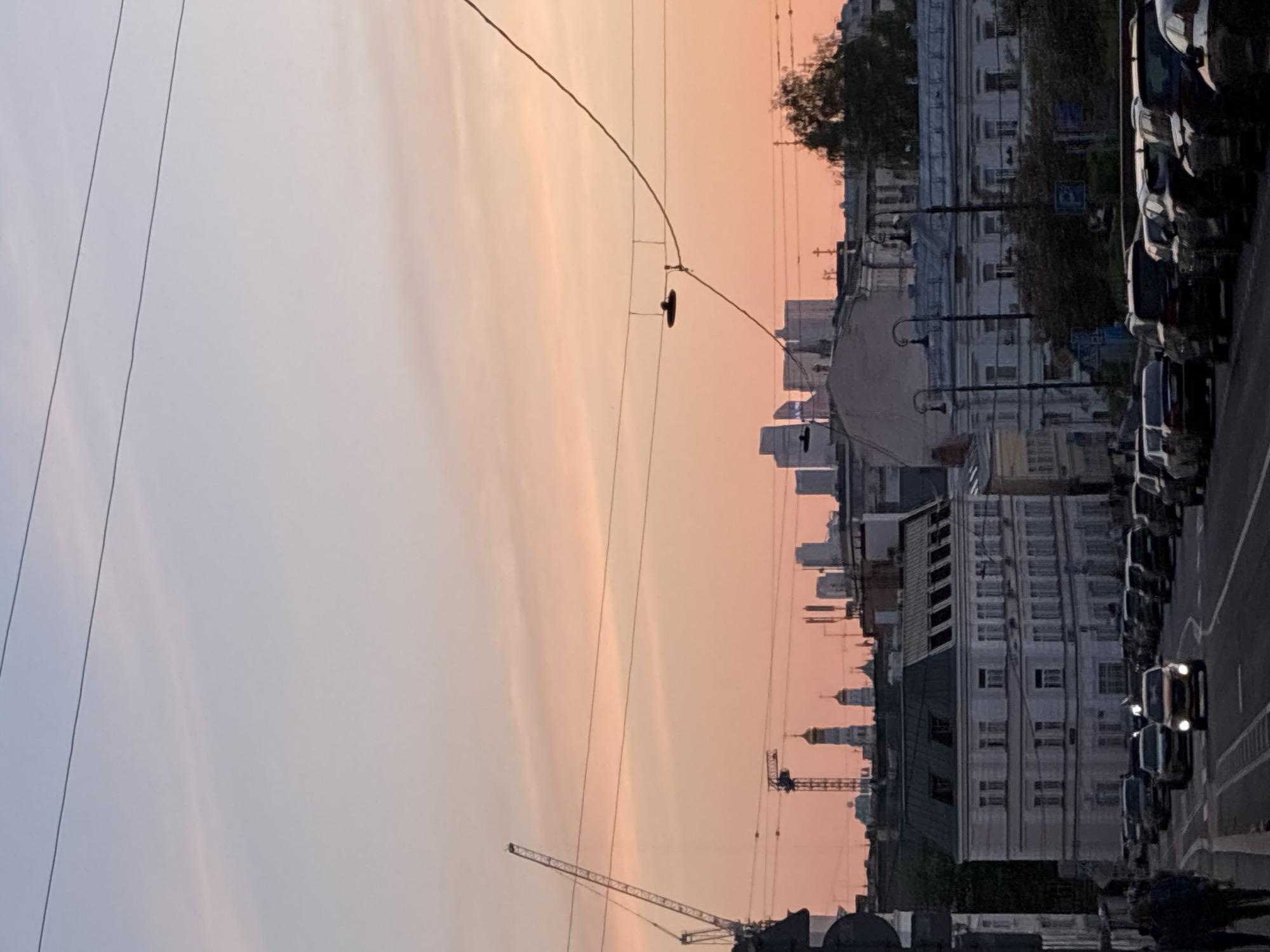 The image shows a street scene in Moscow, Russia, at sunset. A line of parked cars runs along the right side of the frame. Buildings, including some with prominent spires and cupolas, line the street. A construction crane is visible against the sky. Overhead wires crisscross the sky. The sunset casts a pink hue. A car drives along the street.