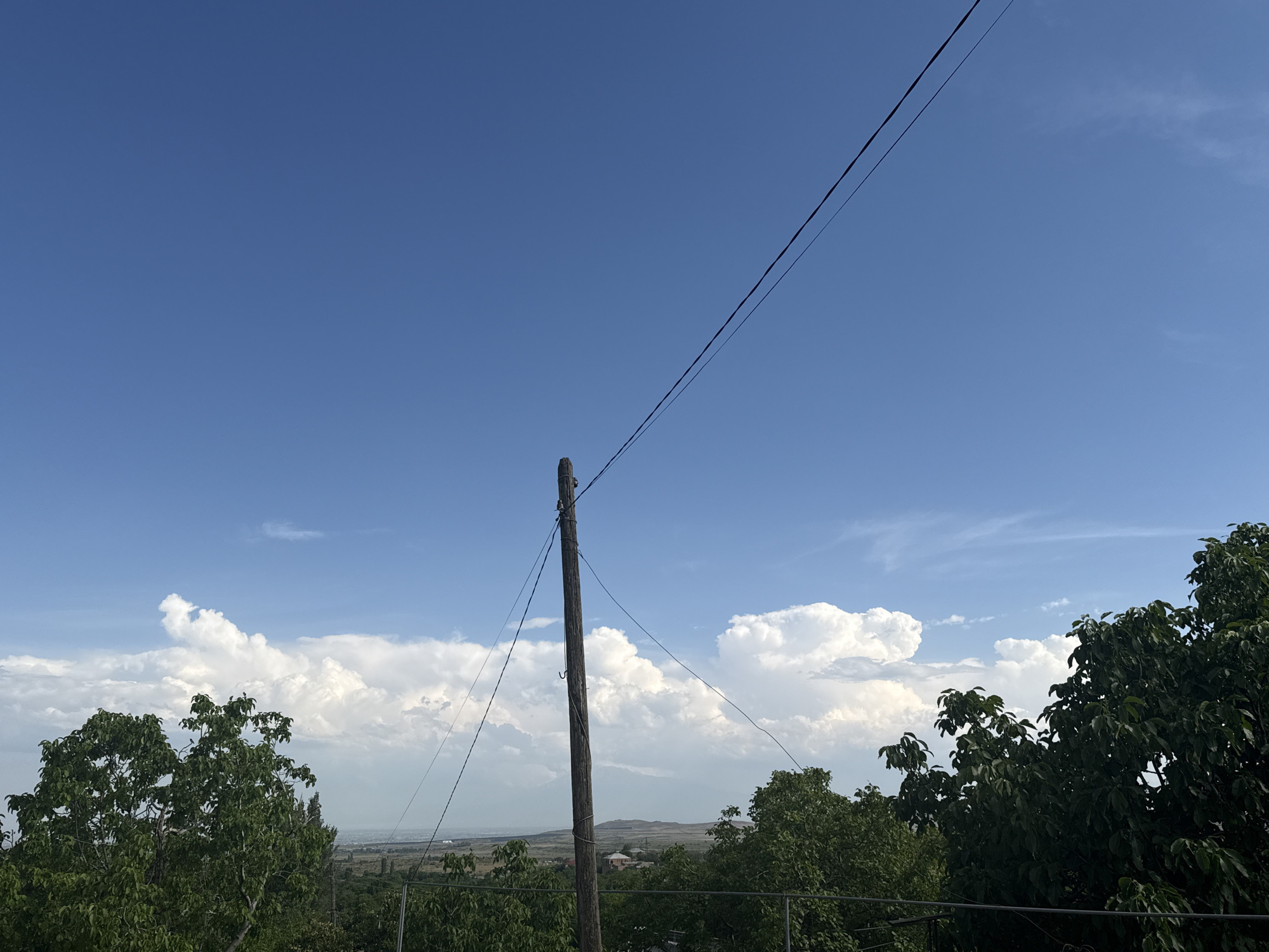 The image depicts a scene dominated by a vast blue sky with scattered white, fluffy clouds. In the foreground, a weathered wooden utility pole stands vertically, supporting dark electrical wires that stretch diagonally across the frame. Lush green trees frame the bottom and right side of the image, suggesting an outdoor, possibly rural or suburban, environment. In the distance, a hazy landscape with rolling hills and a few buildings can be seen under the bright sky. The overall impression is one of a clear, sunny day with a slightly overcast sky.