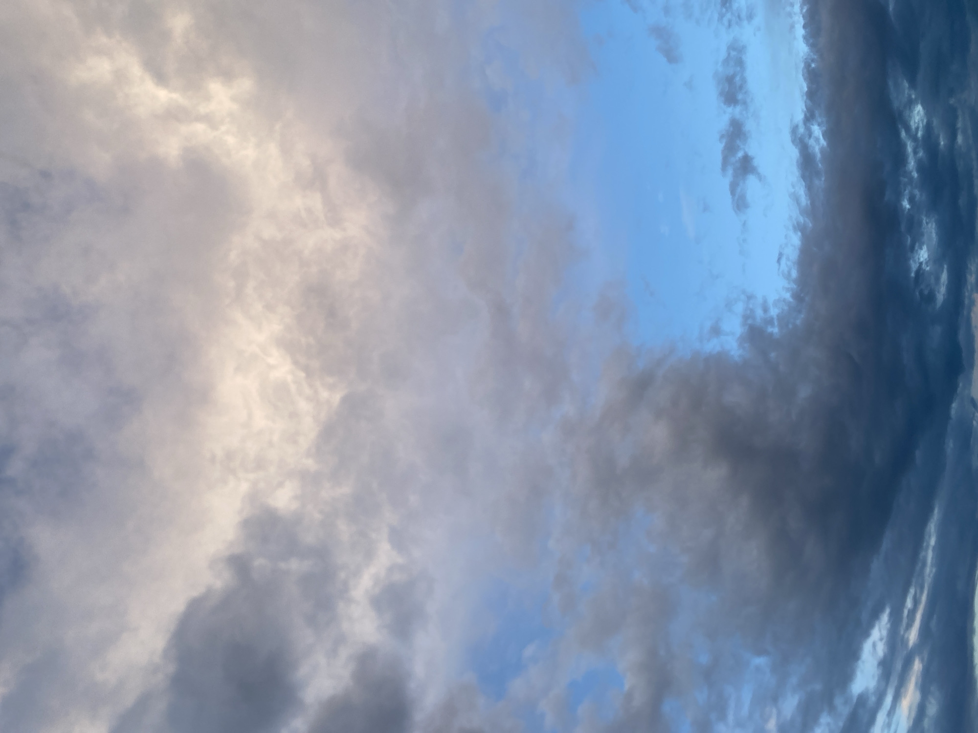 The image is a vertical shot of a cloudy sky. The clouds are varied in color and density, ranging from a bright, sunlit white at the top to dark, ominous gray at the bottom. Patches of clear blue sky are visible in the middle section. The scene appears to be an outdoor environment, possibly captured in the city of Yablunivka, Ukraine, given the context provided. There are no people, subjects, or specific objects discernible in the image beyond the clouds and sky. The environment is atmospheric, characterized by the dynamic interplay of light and shadow within the cloud formations.