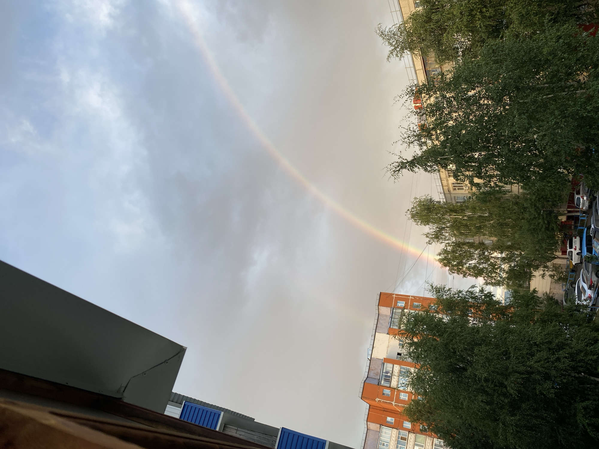 The image captures a double rainbow arcing across a cloudy sky over a residential area in Surgut, Russia. In the foreground, a portion of a building or balcony is visible in the upper left corner. The middle ground is dominated by several multi-story apartment buildings, some with orange and white facades, and lush green trees with dense foliage, possibly birch trees, interspersed between them. In the background, more apartment buildings are visible. The lower portion of the image shows a street with several parked cars. The scene suggests a post-rain or stormy weather condition, typical of a summer day in Surgut.