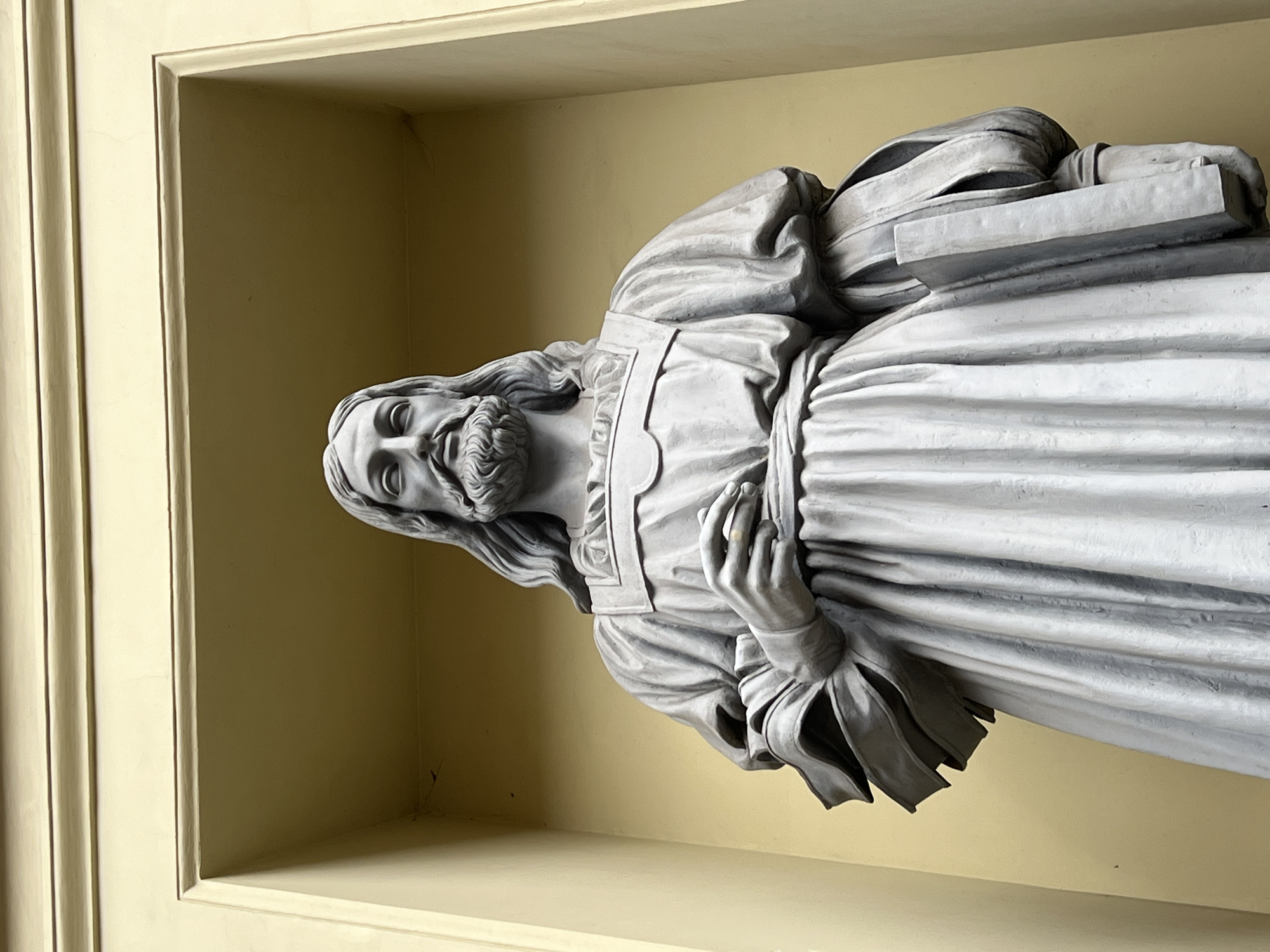 A grey stone statue of Jesus Christ stands in a niche of a pale yellow building. The statue depicts Jesus from the chest up, with long wavy hair, a beard, and a serene expression. He is wearing a flowing robe with a sash around his waist and holds an open book in his left hand. The statue is lit from above, casting shadows on the sculpture. The background is a plain yellow wall within the niche. The building appears to be constructed of light-colored stone or plaster. The architectural style suggests a historical European setting, possibly a church or a public building in Saint Petersburg, Russia, given the similar niche carvings found on many of its neoclassical structures.
