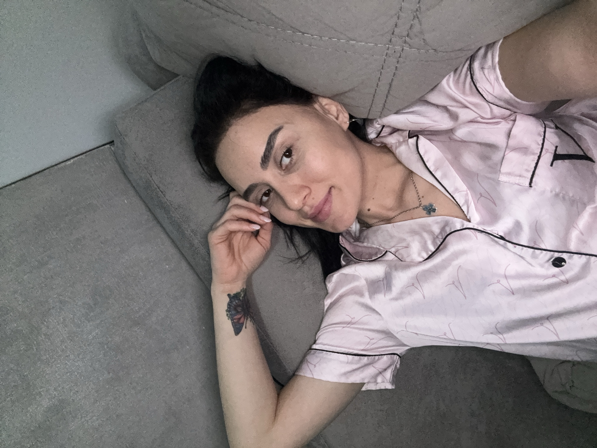 A woman lies on a grey fabric couch. She is fair-skinned with dark hair pulled back, and is wearing light pink satin pajamas with black trim. She is smiling softly at the camera, her left hand near her face. A butterfly tattoo is visible on her left forearm. A silver necklace with a four-leaf clover pendant is around her neck. The location is not identifiable as Tbilisi, Georgia.