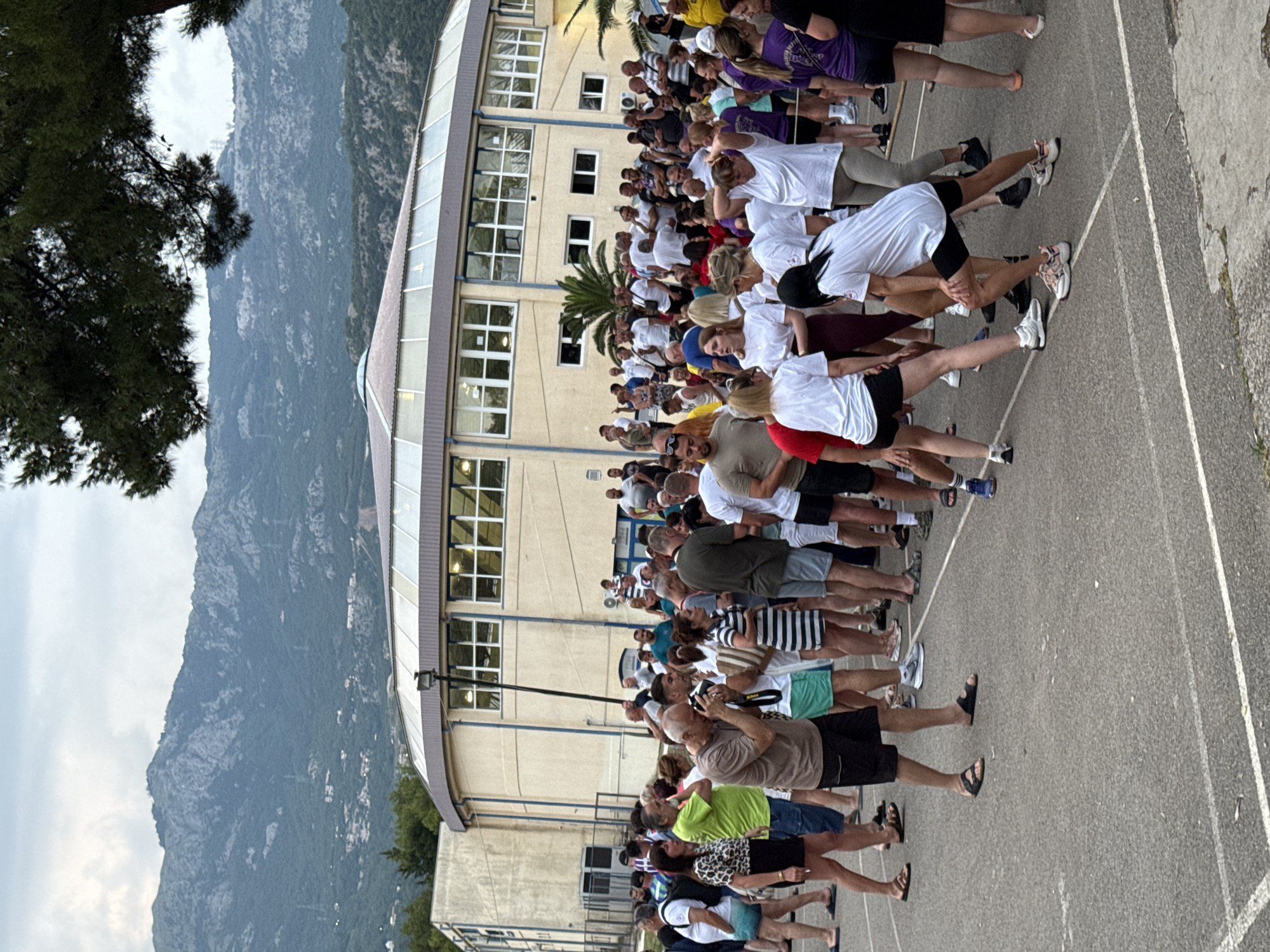 A group of approximately 50 people are standing in rows on an asphalt surface, with a building behind them and a mountainous landscape in the distance. The subjects appear to be engaged in some kind of outdoor activity or class, as they are all facing the same direction and many are in a similar pose, some with arms outstretched or bent. The people are diverse in age and gender, wearing casual clothing such as t-shirts, shorts, and athletic wear.
The building behind the group has a light-colored facade with many windows and appears to be a school or community center. Palm trees are visible near the building. The mountainous landscape in the background suggests a Mediterranean or coastal setting. The time of day appears to be late afternoon or early evening, based on the soft lighting and the shadows. The weather seems to be overcast.
There is no visible text in the image. The overall mood of the scene is one of communal activity and possibly exercise or training.