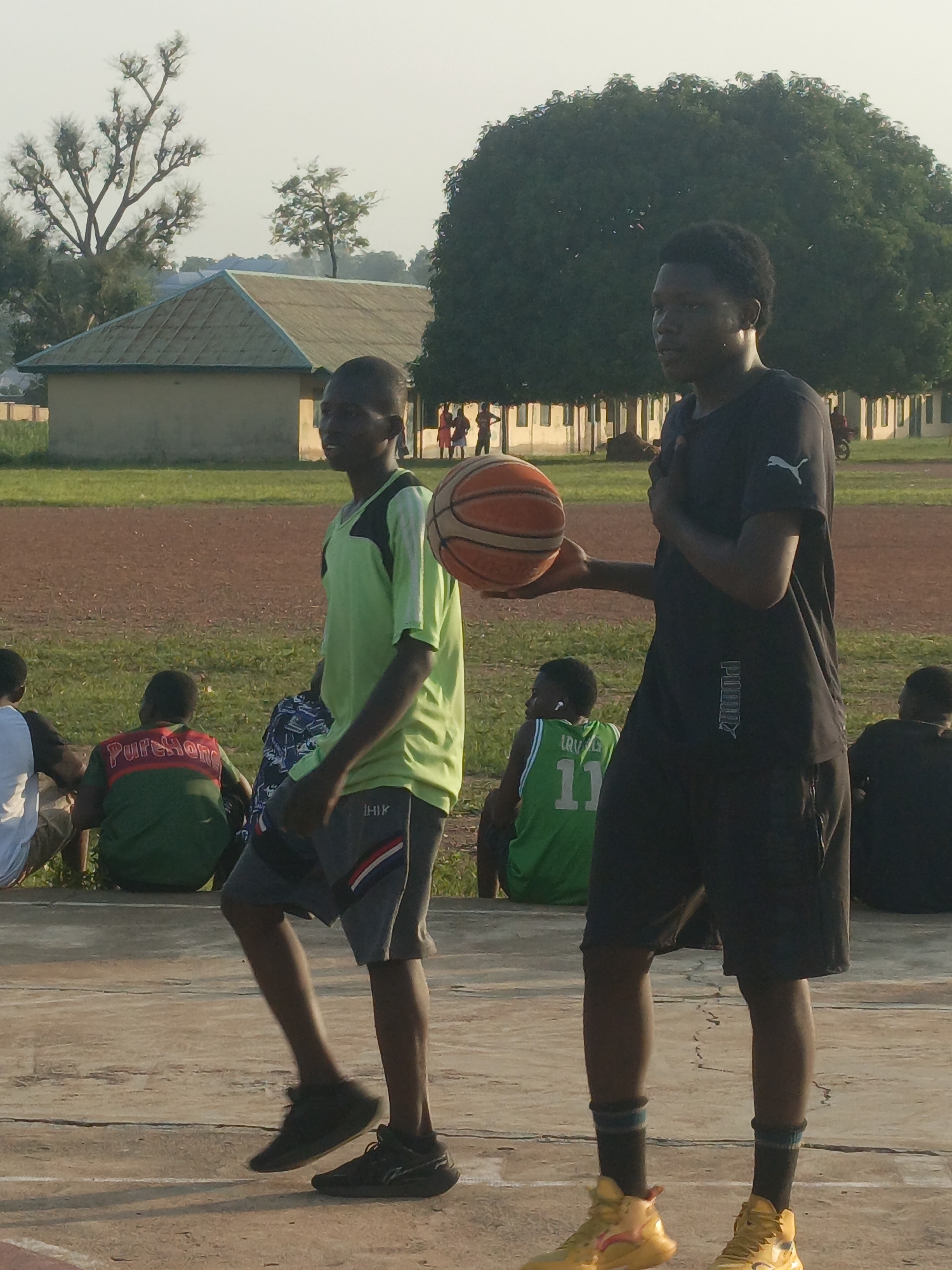 In Akwanga, Nigeria, two young men are positioned on a basketball court, with one holding a basketball. The individual on the right, wearing a black Puma t-shirt and black shorts, has his right hand holding the basketball and his left hand on his chest. He is wearing yellow athletic shoes. To his left, another young man in a lime green t-shirt and grey shorts is walking with his hands behind his back. He is wearing black athletic shoes. Several other individuals are visible in the background, some sitting on the ground. The setting includes a dirt track, a grassy field, and buildings with tiled roofs, suggesting an educational institution or community sports facility. A large tree with dense foliage dominates the background. The sky is overcast, indicating daylight.