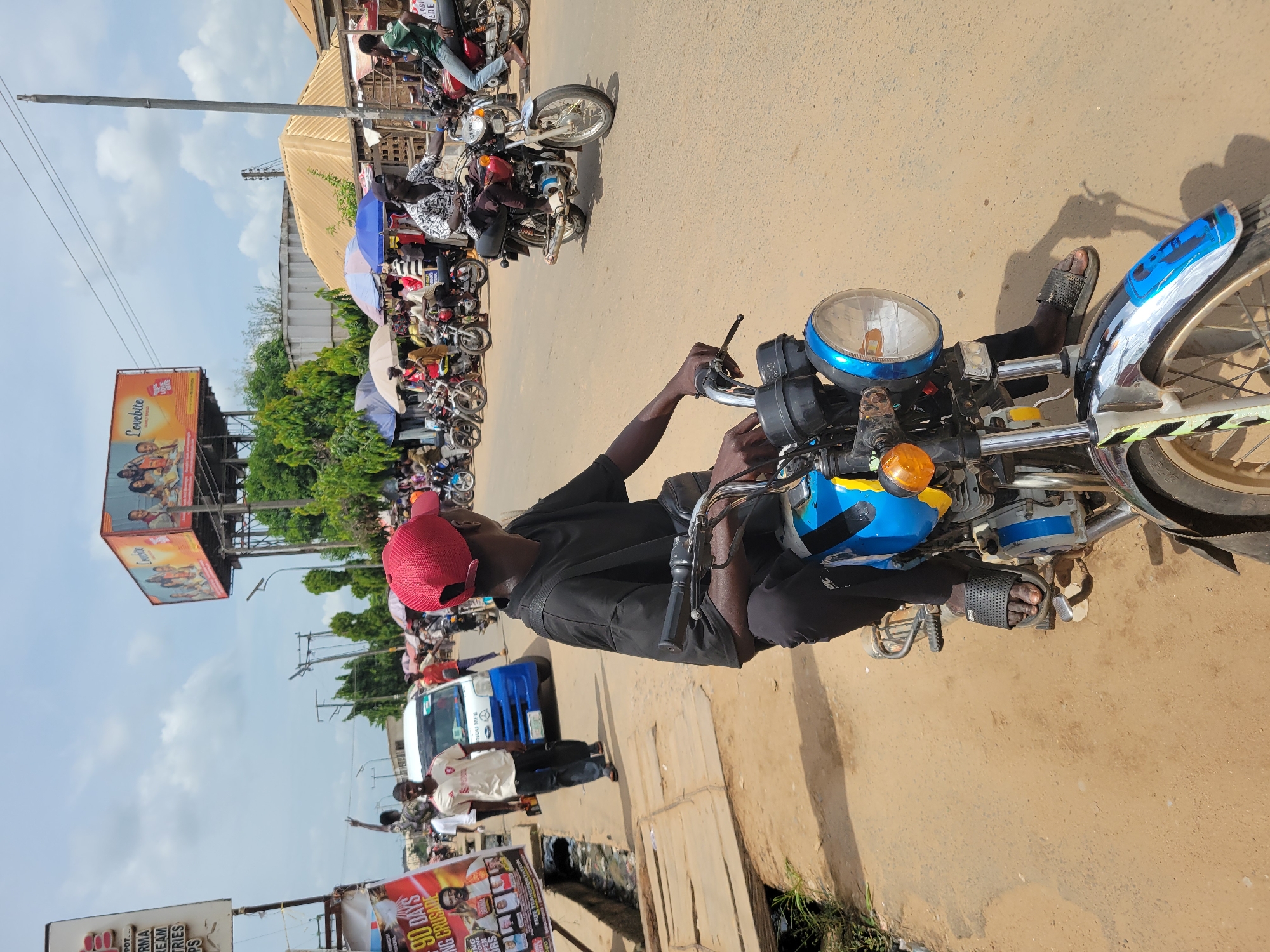 The image shows a man sitting on a blue motorcycle in the city of Egbagu, Nigeria. He is wearing a black shirt, black pants, and a red cap. His feet are clad in sandals. He appears to be adjusting something on the motorcycle's handlebars. The street scene includes other motorcycles parked nearby, some with passengers. There is a white and blue van on the side of the road, and several people are standing in the vicinity. A billboard advertising "Lovebite" snacks is visible in the background, along with political posters displayed near the foreground. The area appears to be an urban environment, possibly a roadside market or a busy intersection.