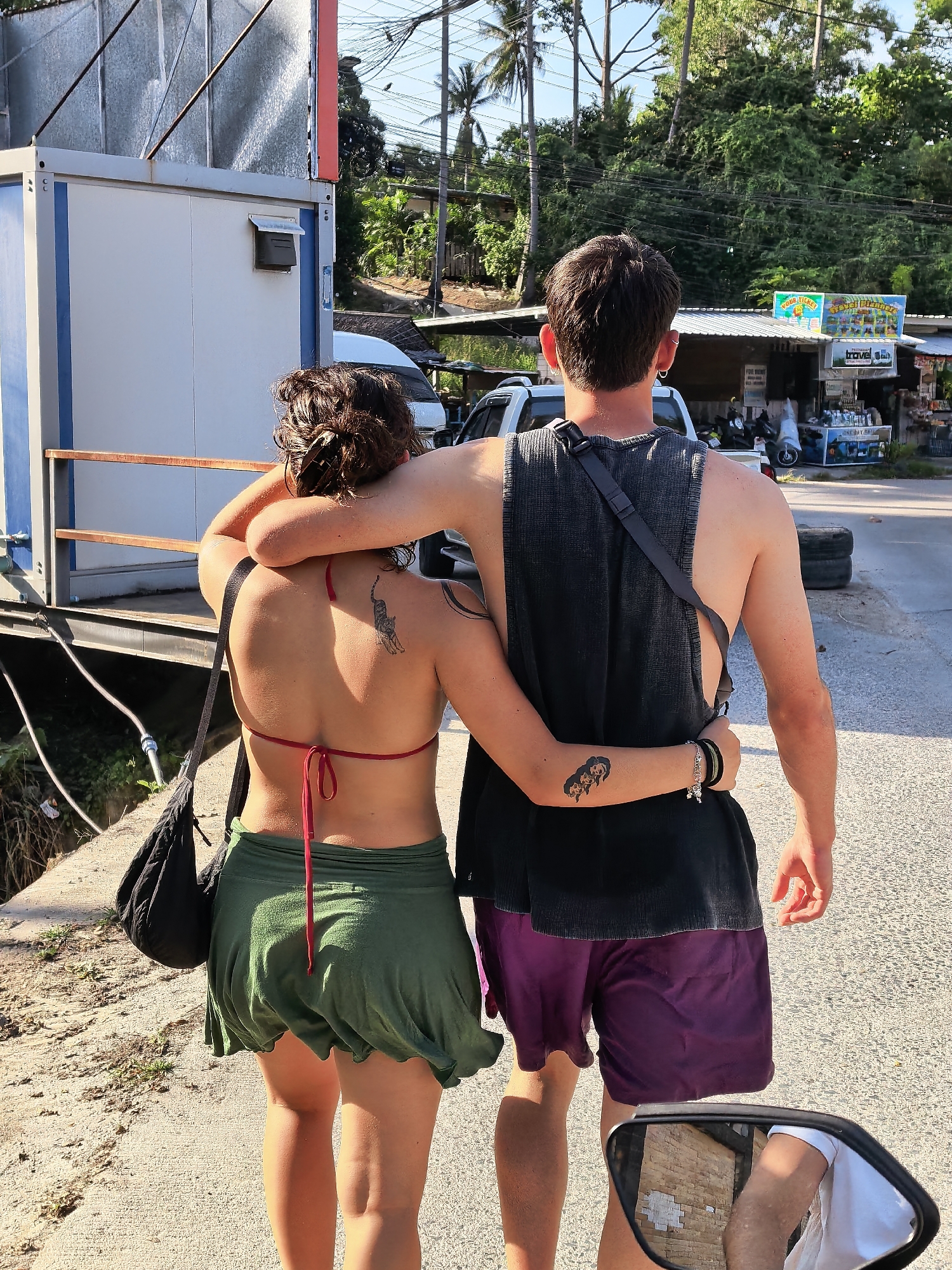 In the city of Ko Samui District, Thailand, a young couple walks arm-in-arm down a paved street. The woman, facing away from the camera, wears a red bikini top and a green, flowy skirt. She has a black tattoo of a cat on her upper back. The man, also facing away, wears a dark grey sleeveless t-shirt and purple shorts. He has a tattoo of three dog heads on his left forearm and a black strap across his chest. A black bag is slung over the woman's shoulder. In the background, a white portable building stands to the left, and a row of small shops with signs, including one advertising "TRAVEL TICKETS," are visible to the right. Palm trees and dense foliage frame the scene under a bright sky. A white SUV is parked on the right side of the street. The image is captured from a slightly elevated perspective, possibly from the back of a motorcycle as suggested by the rearview mirror reflecting a person in a white t-shirt and a brick building.