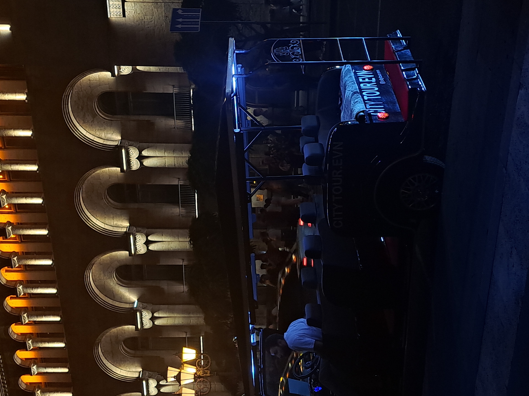 At night in Yerevan, Armenia, a "City Tour EVN" passenger vehicle with the driver at the wheel is stopped on the street next to a building decorated with illuminated arches. Several passengers sit in the back of the vehicle.