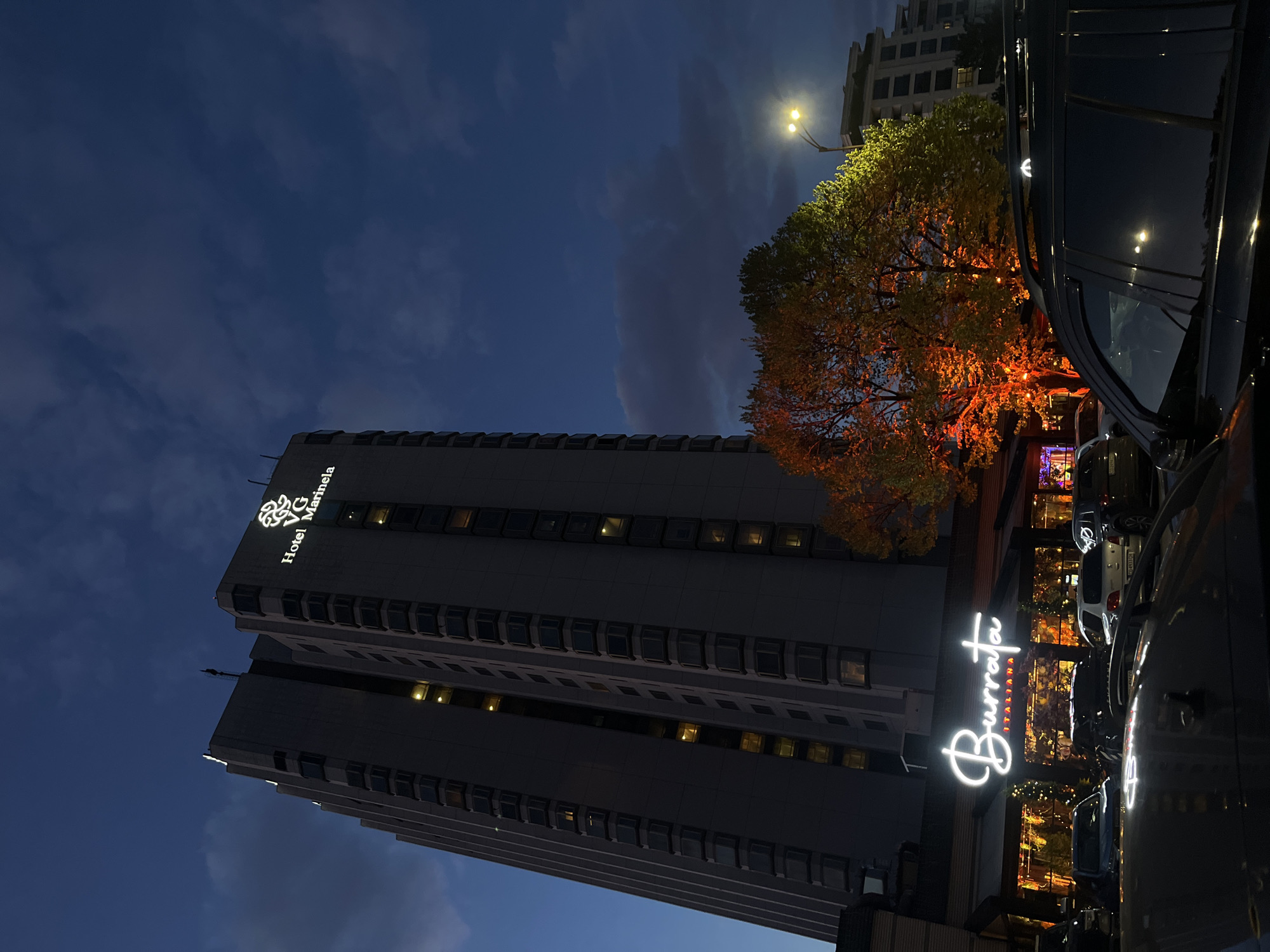 The image depicts the Hotel Marinela in Sofia, Bulgaria, at night. The hotel is a tall building with a sign bearing its name and logo. Next to the hotel is the "Burrata" Italian restaurant, whose illuminated signage is visible. A tree with leaves illuminated in orange stands between the hotel and the restaurant. Several cars are parked along the street. The sky is dark with clouds.