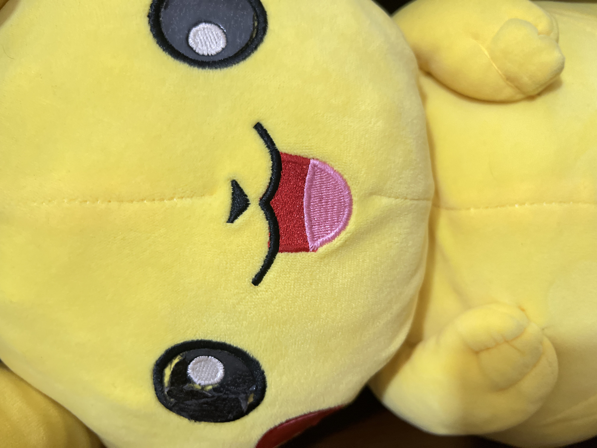 The scene, situated in Jiulongpo District, China, presents a detailed, close-up, frontal view of a yellow plush toy, prominently resembling the character Pikachu from the Pokémon franchise. The toy occupies the majority of the vertical frame, showcasing its embroidered facial features. It has large, round black eyes, each with a distinct white circular highlight. A small, black, triangular nose is centered above a wide, open-mouthed smile. The mouth is outlined in black and reveals a textured red upper tongue and a textured pink lower tongue playfully extended outwards. A red circular cheek marking is partially visible on the left side of its face. The plush material has a soft, slightly textured appearance. At the bottom of the frame, parts of the toy's yellow, rounded body and two small, stubby arms are visible. The background is uniformly dark and out of focus, bringing the plush toy into sharp relief.