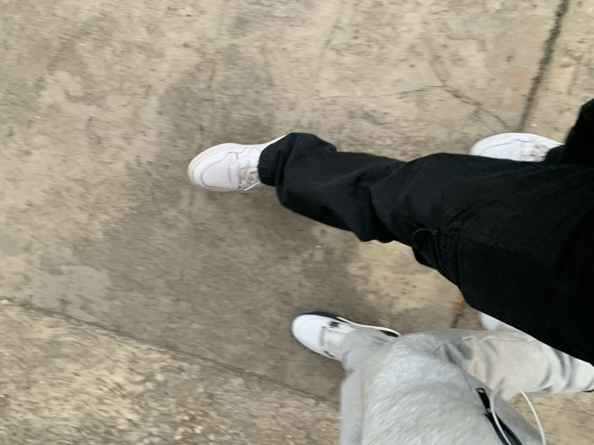 This image, taken from an overhead perspective, captures the lower halves of two individuals walking or standing on a concrete surface in Pampatar, Venezuela.
The subject on the left wears loose-fitting black pants and white sneakers with visible laces. Their leg is extended forward, suggesting movement. The subject on the right wears light grey or white sweatpants, which appear ribbed or textured, and white sneakers with dark blue or black accents near the ankle/heel. A white cable, possibly from earphones, dangles near their leg.
The ground is a textured, light grey concrete or asphalt, showing various shades of grey, indicating wear or natural variation. The overall scene appears somewhat blurry, particularly towards the edges, which could be due to motion blur, a shallow depth of field, or the camera's angle of capture. The lighting is even, suggesting an overcast day or possibly late afternoon/early morning, as there are no distinct shadows. No visible text is present in the image.
