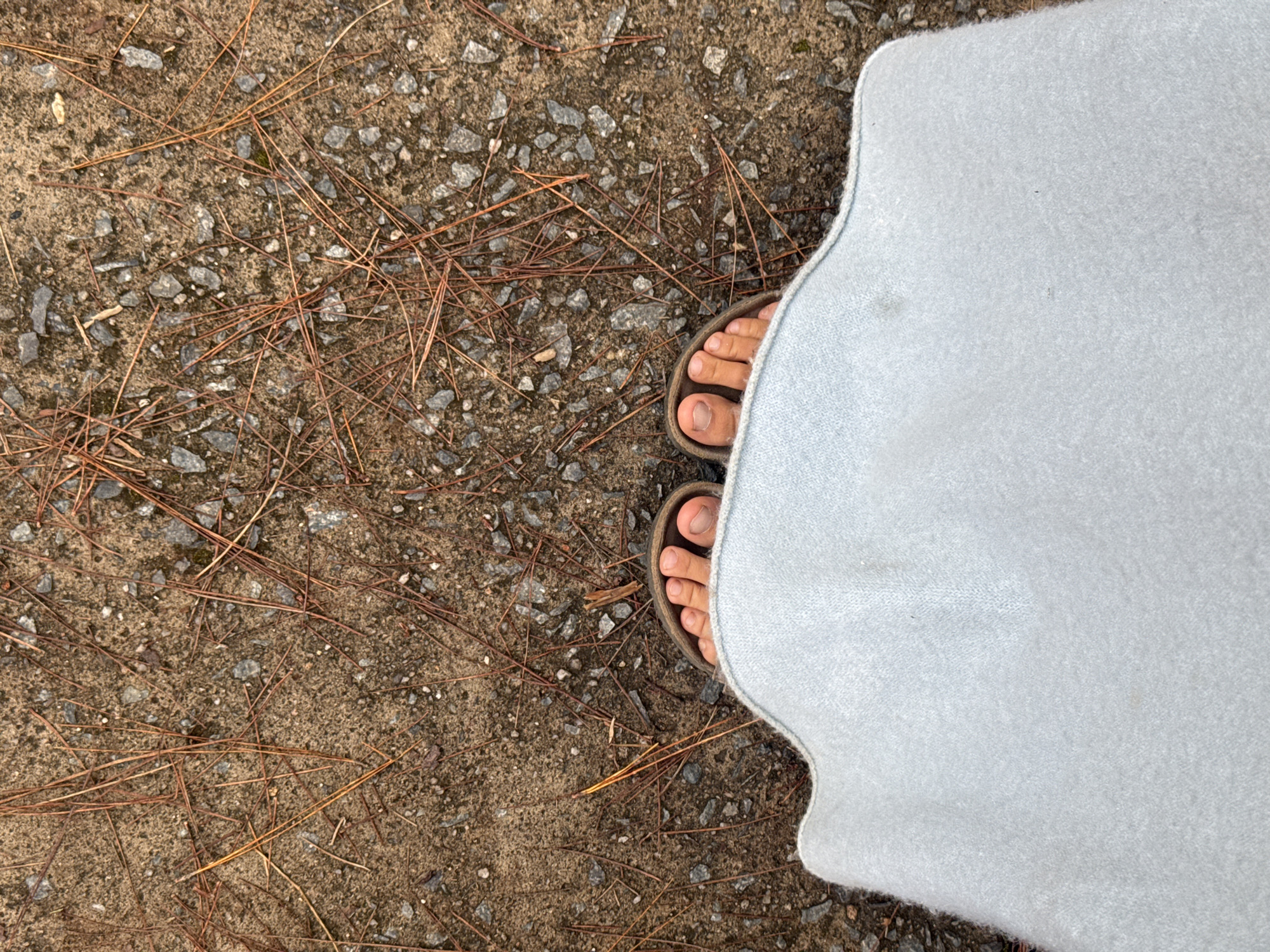 This image shows a close-up, top-down view of a person's feet wearing sandals, peeking out from under a light blue blanket. The feet are standing on a path covered with dirt, small stones, and pine needles. The ground appears to be a natural surface, possibly a trail or unpaved area. The lighting suggests it is daytime. No other subjects or specific landmarks are visible. The location in Đạ Cháy, Vietnam, is not discernible from the visual information present in the image itself.